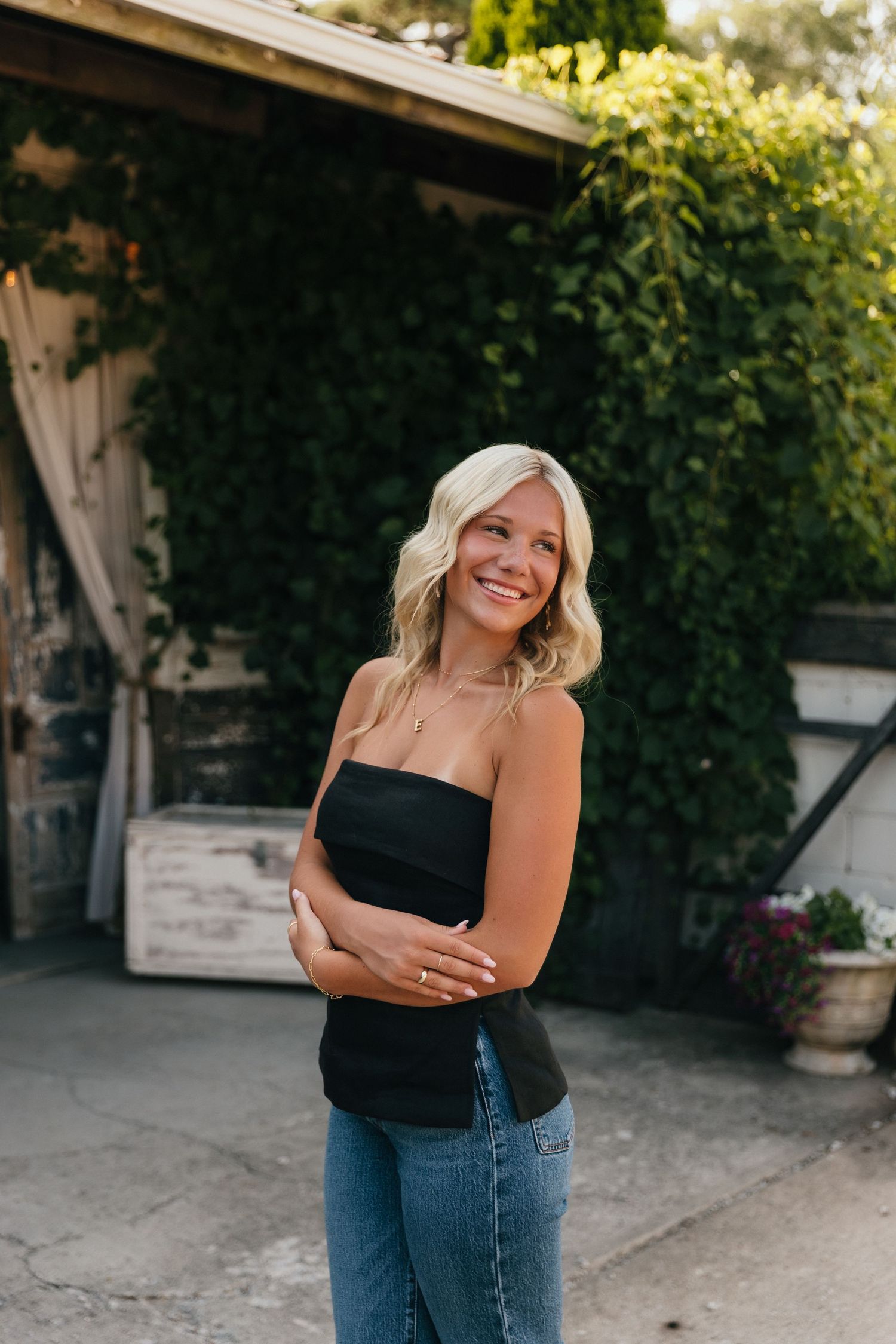 Smiling blonde woman in black tube top and jeans standing outdoors near ivy-covered wall.