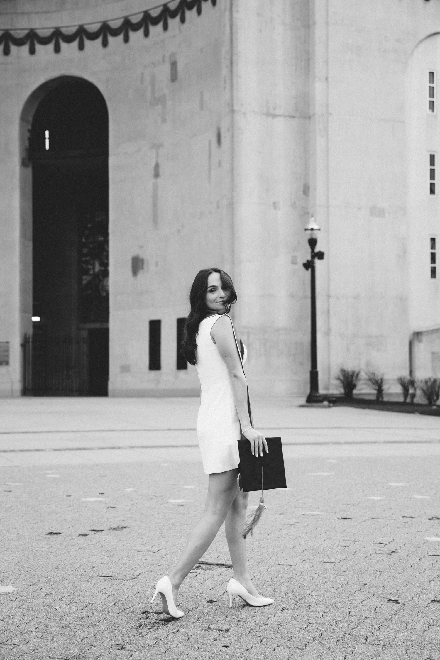 Black and white photo of elegant woman in white dress and heels walking near grand building.