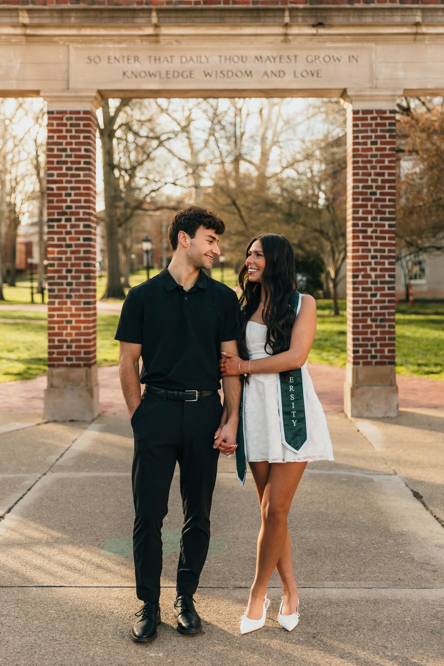 A couple holds hands smiling at each other under a brick arch at a college campus graduation photo.