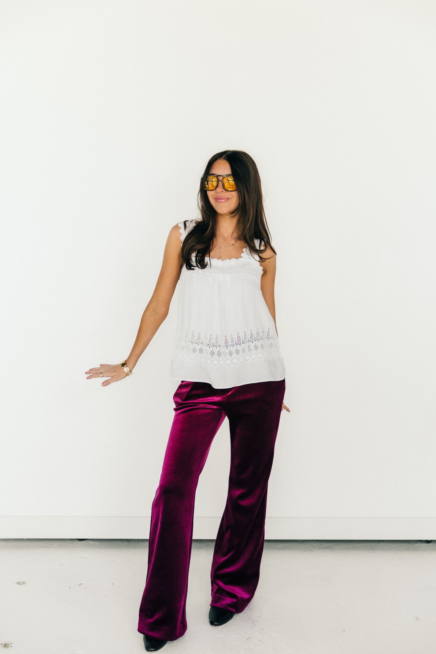 Woman in white lace top, burgundy velvet wide-leg pants, and orange sunglasses posing against a white wall.