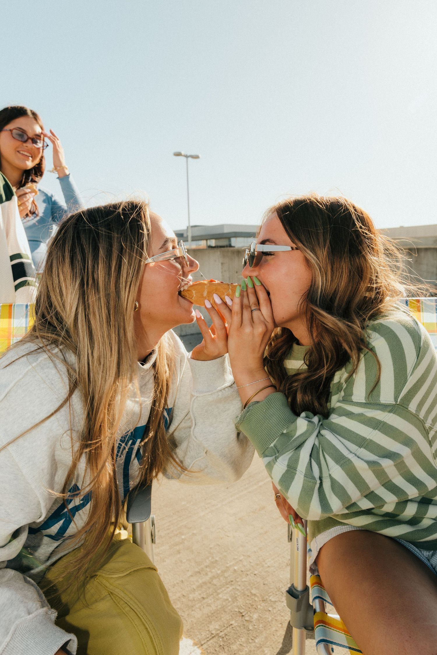 Two young women sharing a slice of pizza outdoors on a sunny day, with a friend smiling in the background.