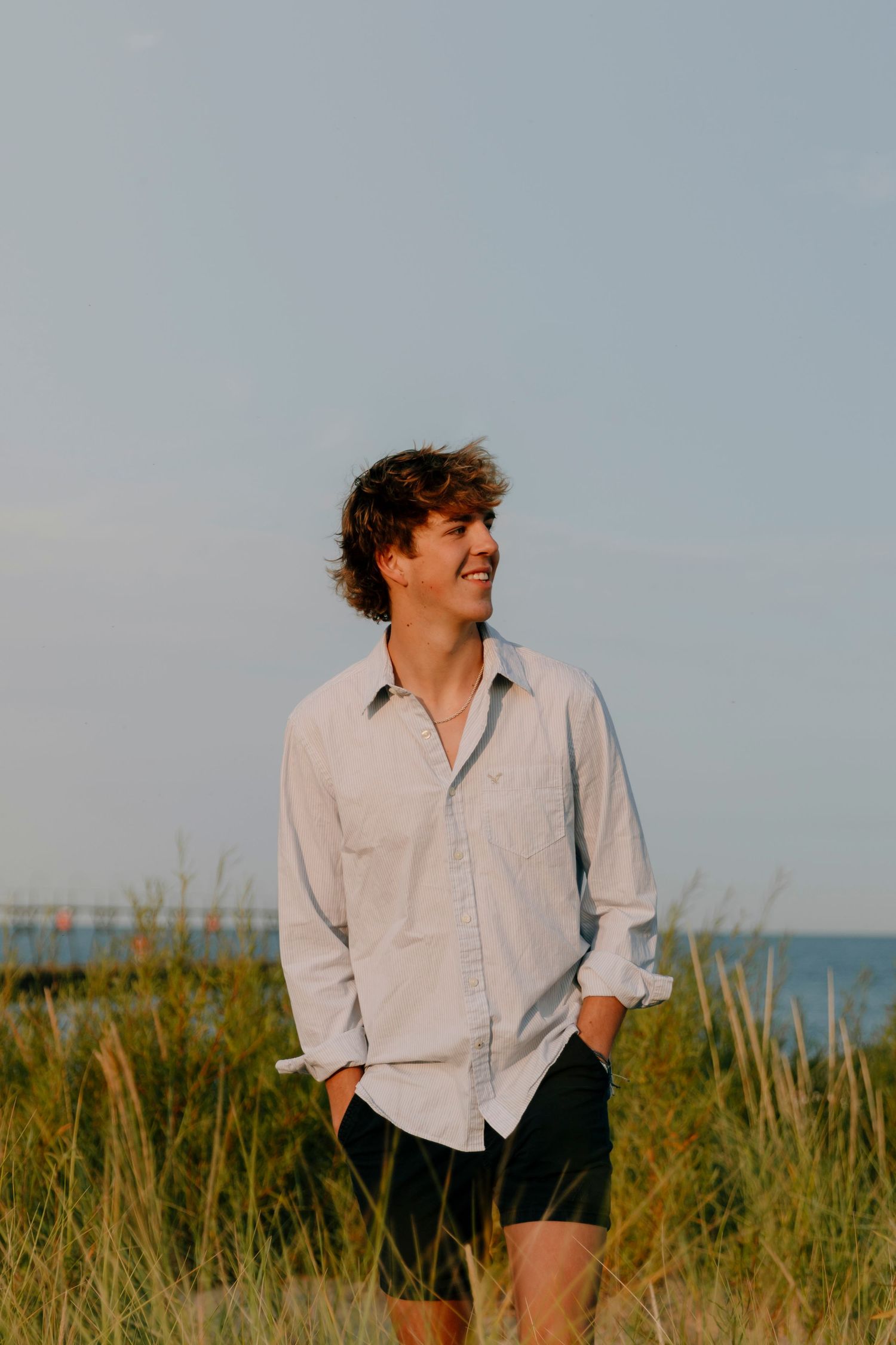A person in a white button-down shirt stands thoughtfully in tall beach grass with the ocean visible in the background.