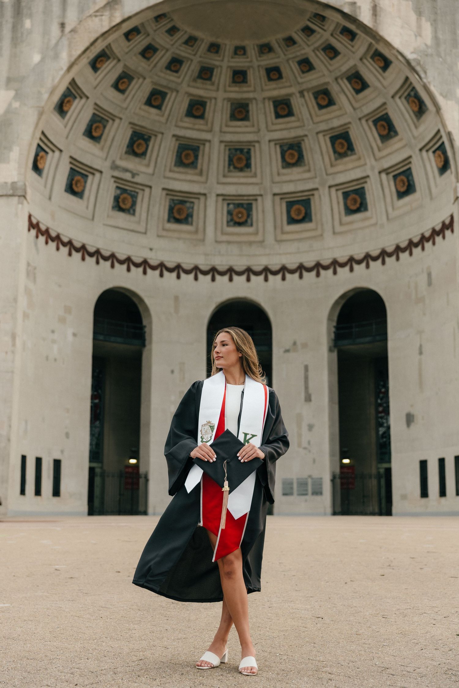 Female graduate in black robe and red sash posing under ornate domed building architecture.