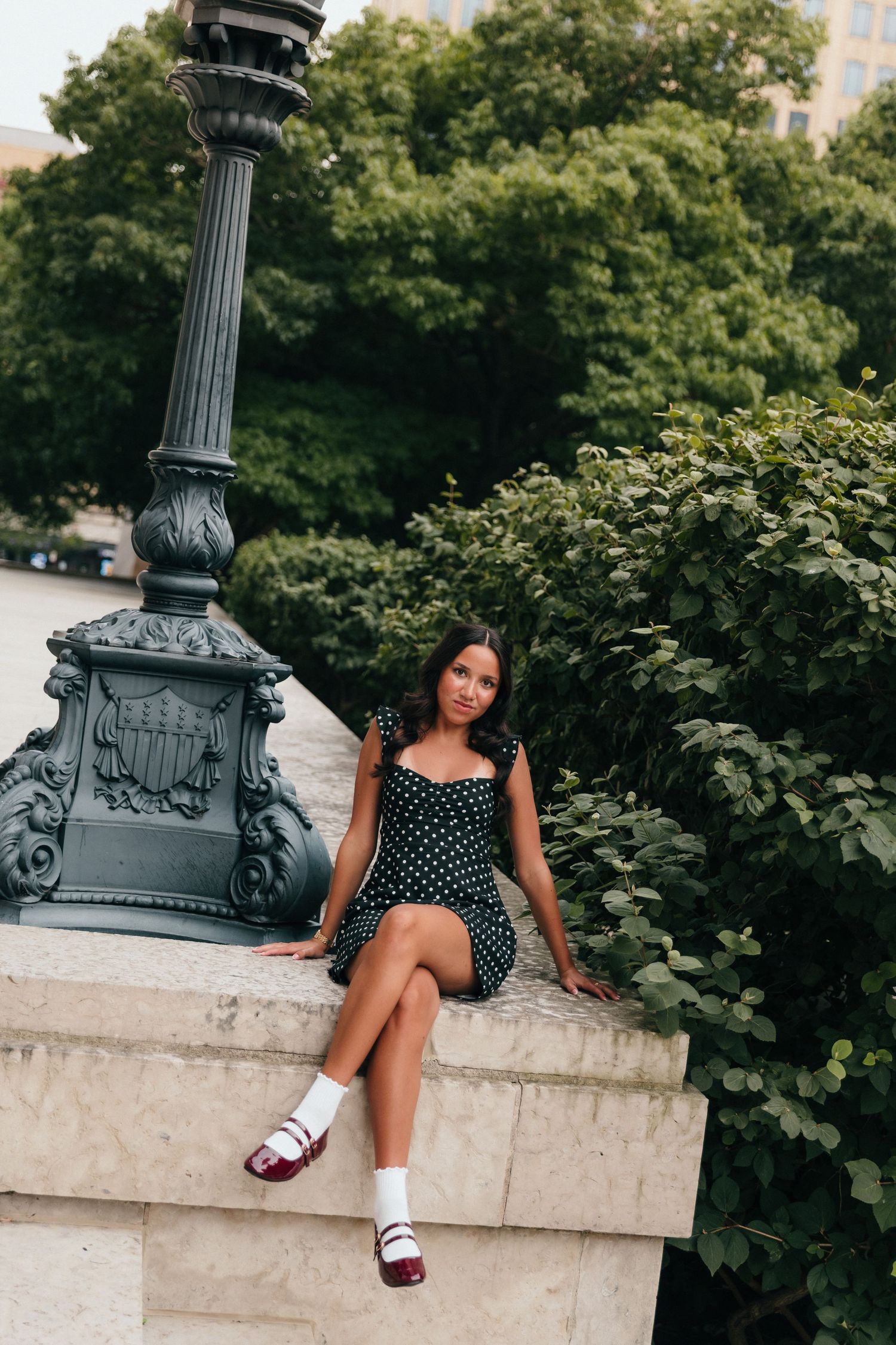 Young woman in polka dot dress and loafers sits on stone ledge near ornate lamp post with green trees behind.