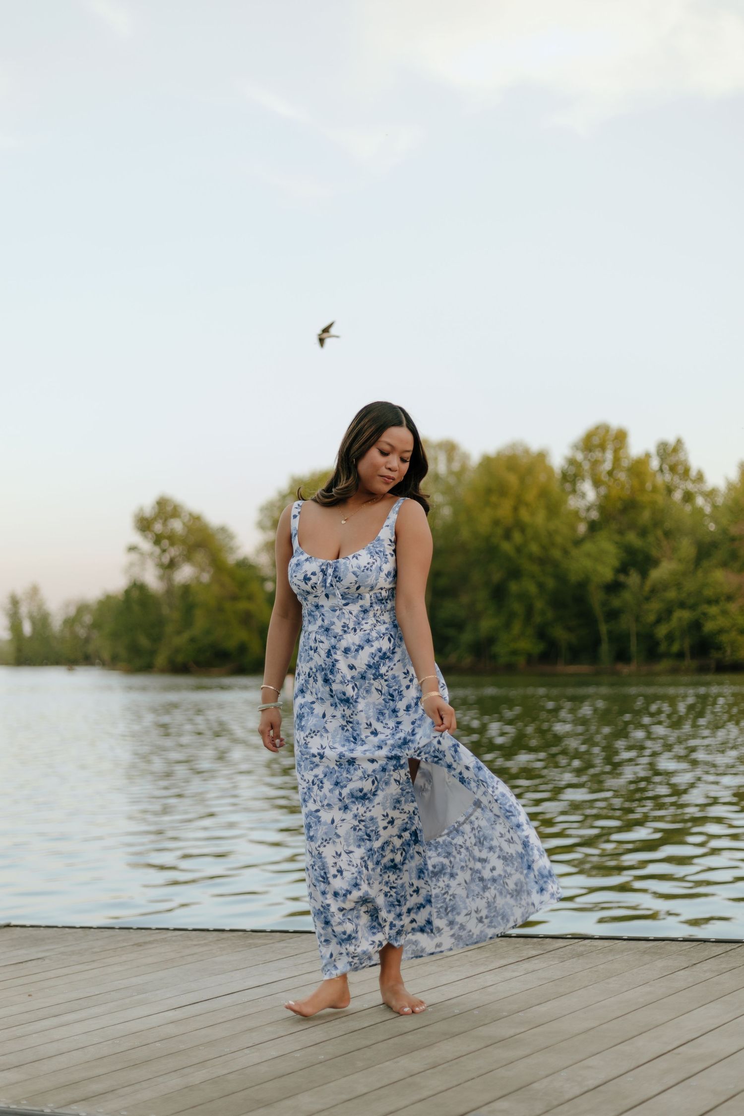 Woman in blue floral dress posing on wooden dock by calm lake.