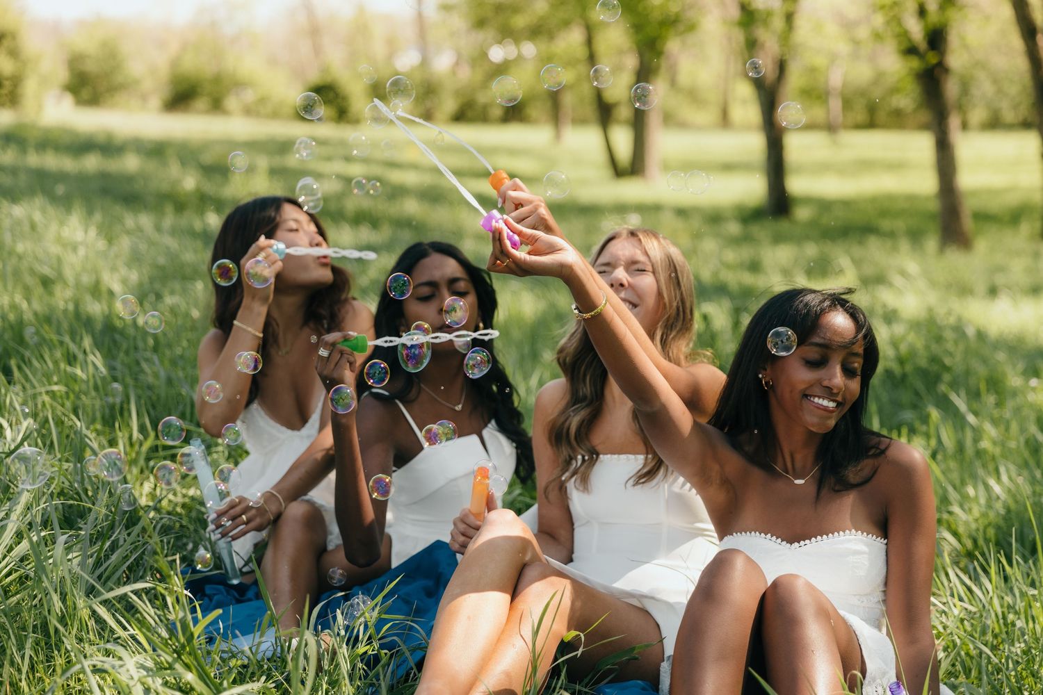 Four diverse women sitting on grass in a park, joyfully blowing bubbles together on a sunny day.
