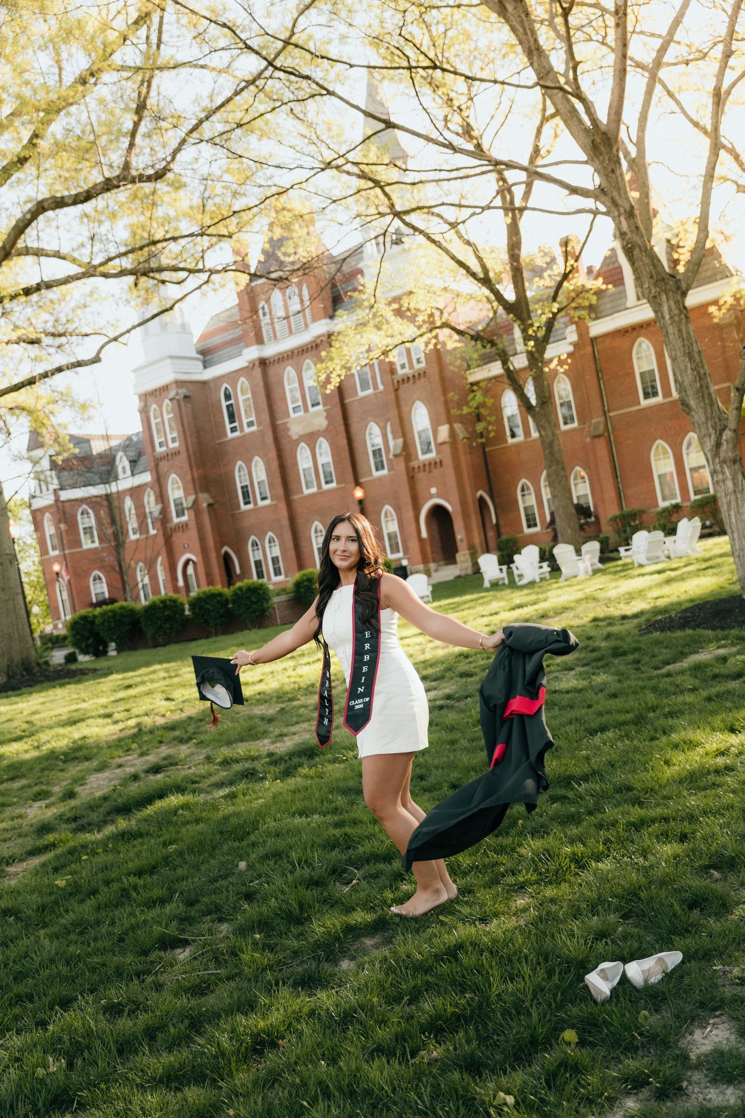 A joyful graduate in a white dress tosses her cap and gown on a sunny college campus lawn with a red brick building.