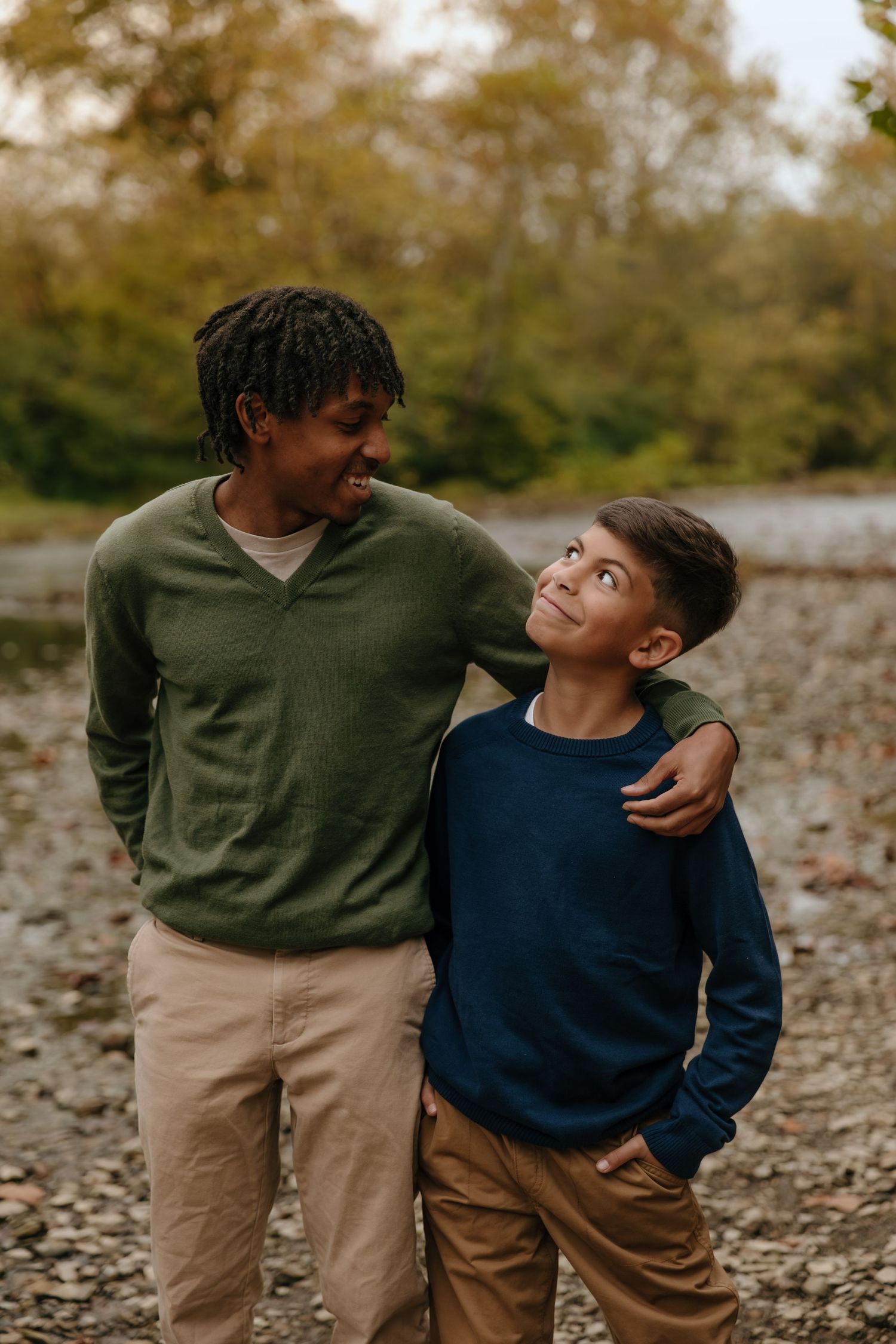 A man and boy in green and blue sweaters share a warm moment outdoors, smiling at each other near a rocky area.