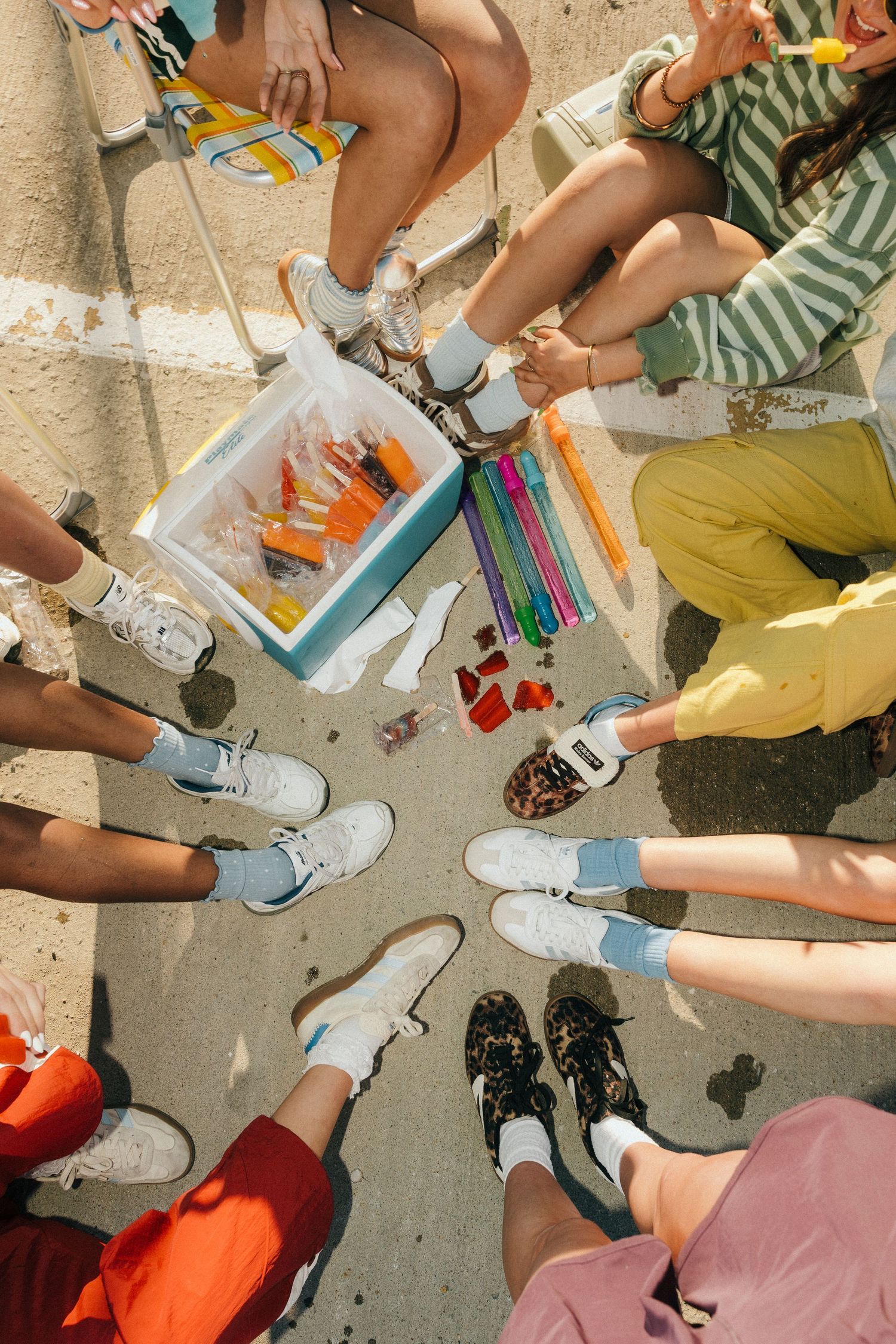 Group of friends sitting in a circle outdoors with sneakers visible and a cooler filled with colorful drinks in center.