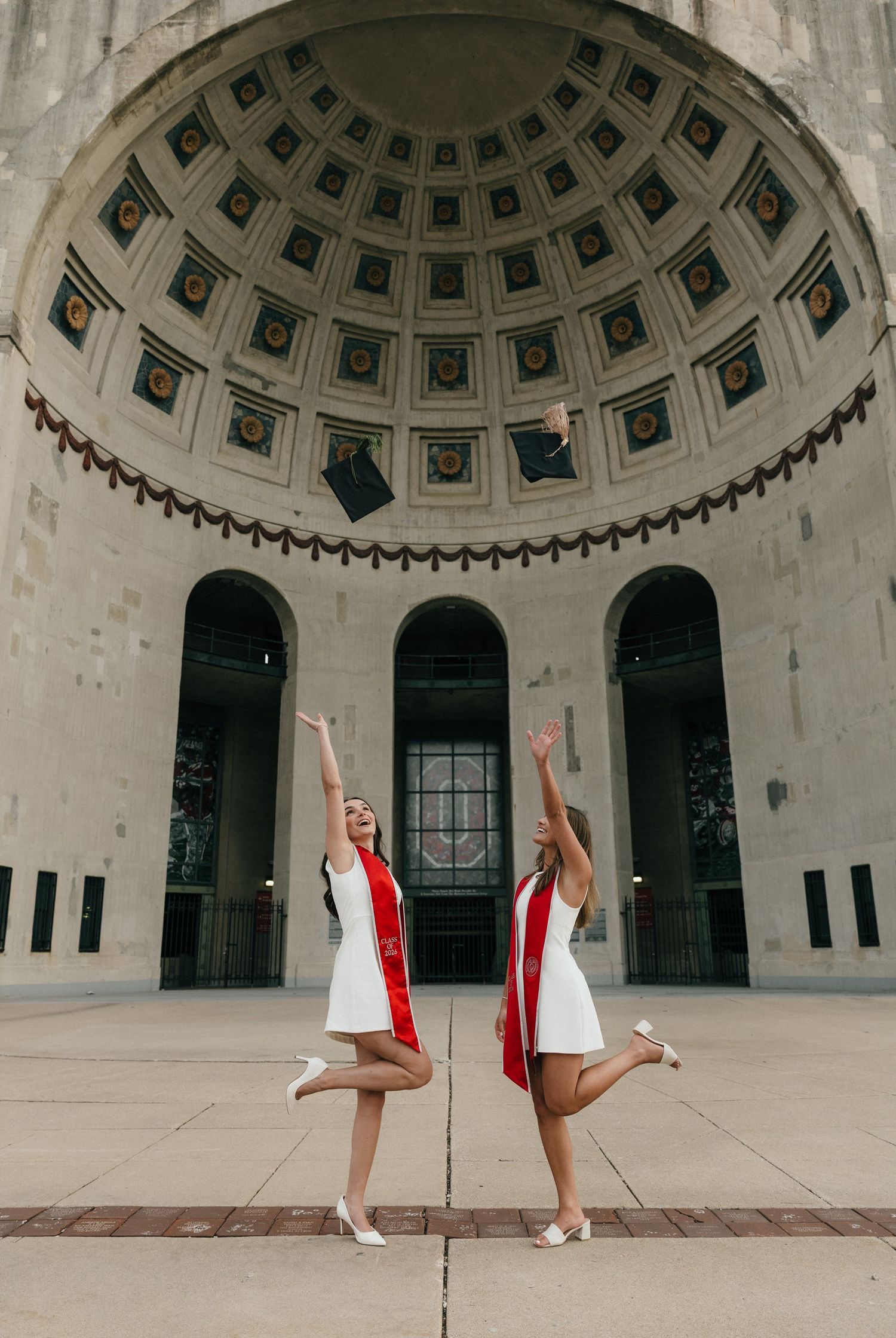 Two graduates in white dresses and red stoles toss caps in air beneath ornate university rotunda.