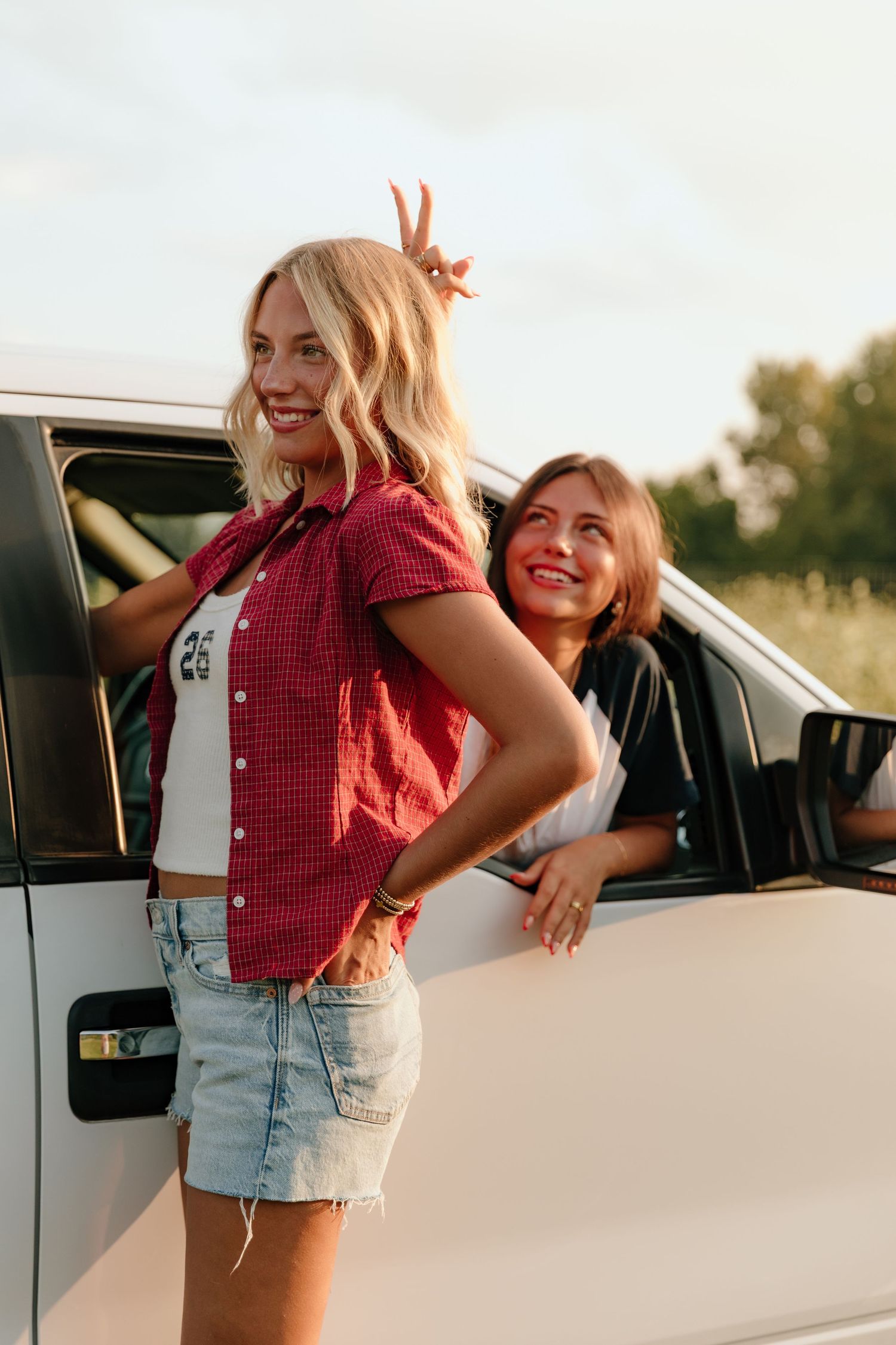 Two young women smiling near a white car outdoors, one making bunny ears behind the other.