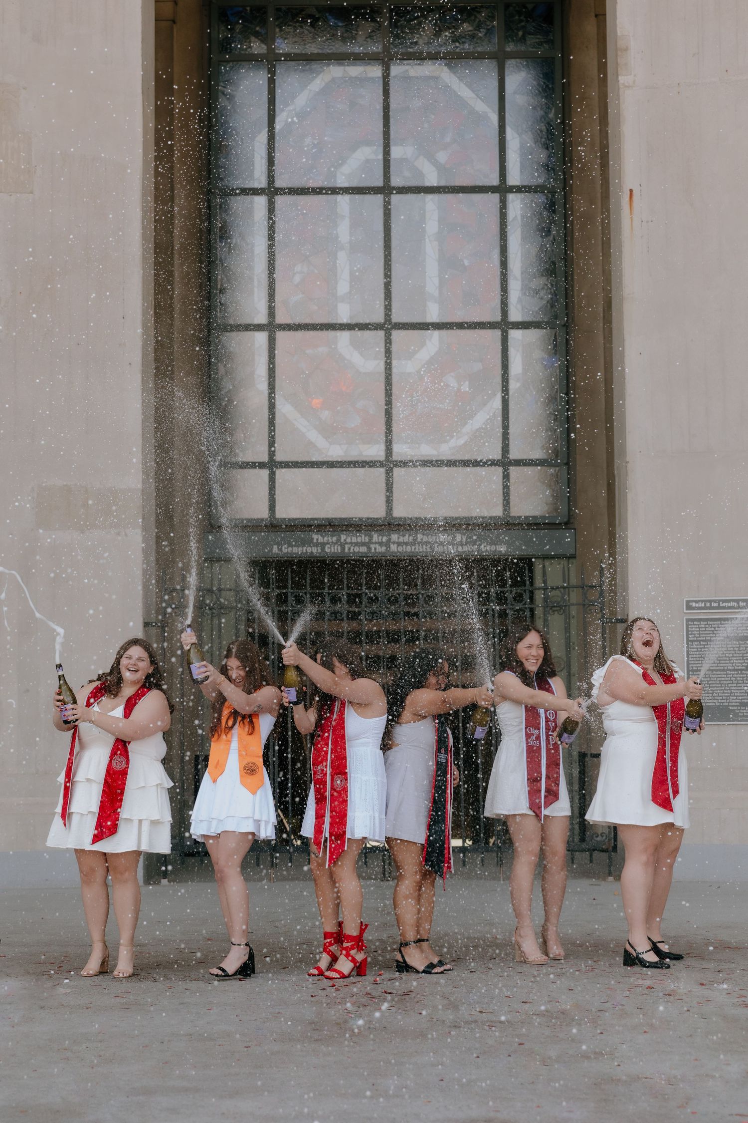 A line of ballet dancers in white and red outfits pose against a gray wall with large windows.