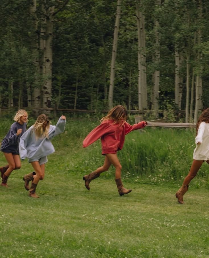 Four girls running and playing in a lush green meadow surrounded by tall aspen trees wearing cozy oversized sweaters.