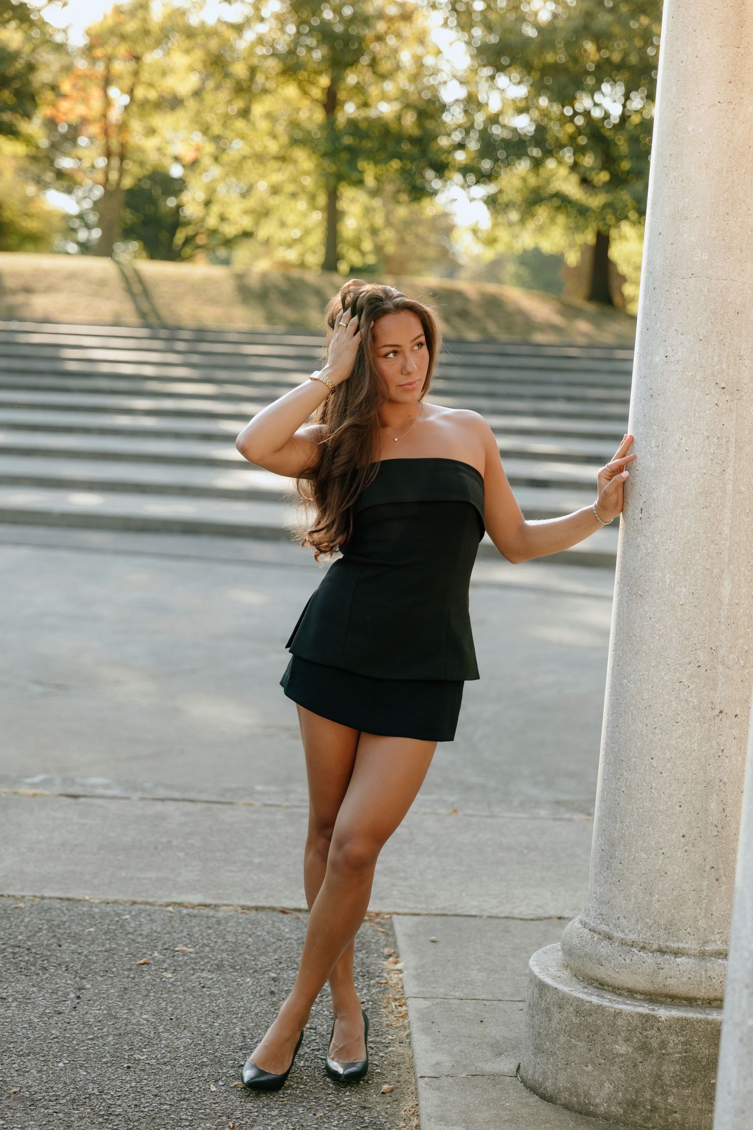 A woman in a black strapless dress poses elegantly against a white stone column in a sunlit outdoor setting.