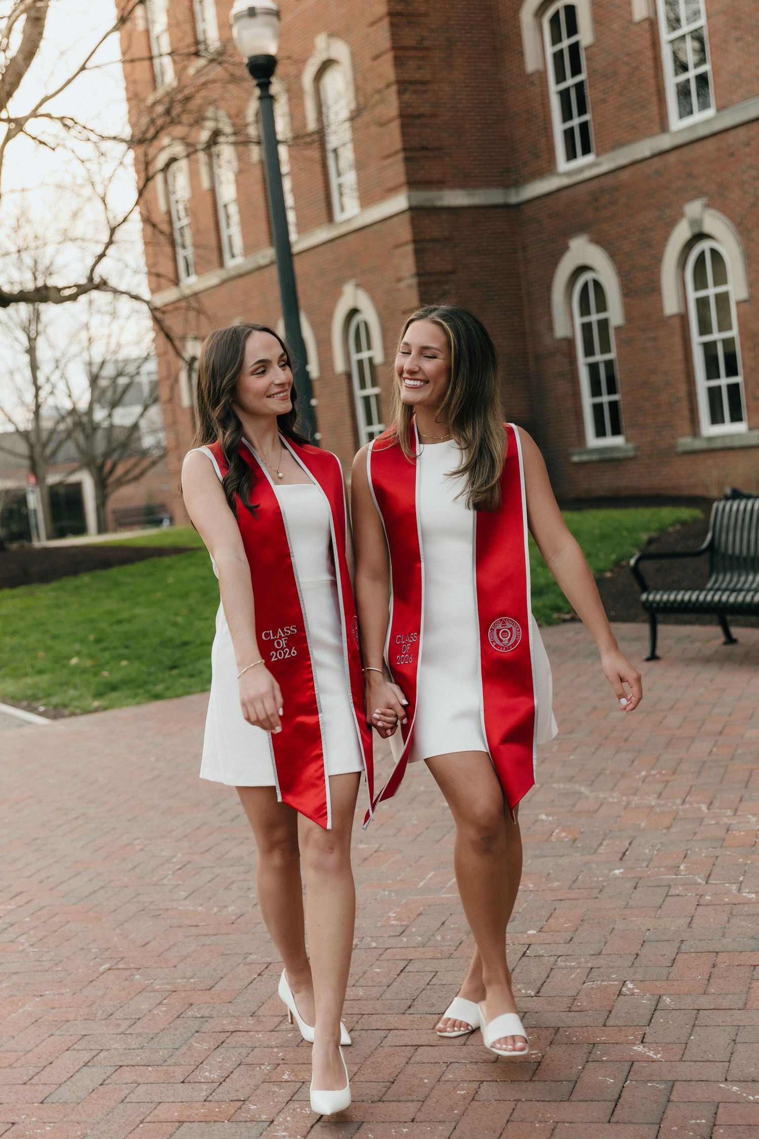 Two graduates in white dresses and red stoles walk hand-in-hand on university brick path.