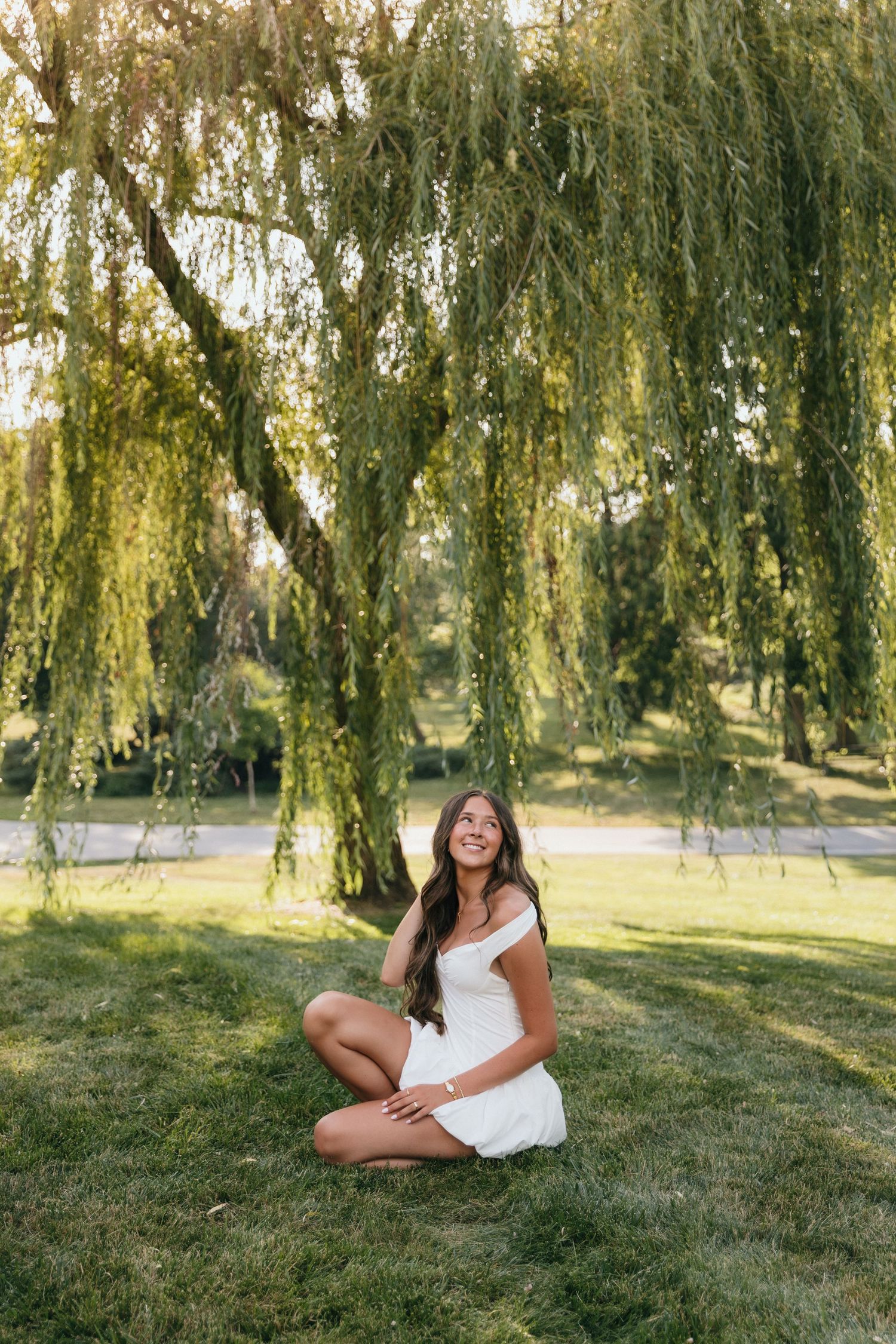 Young woman in white dress sitting cross-legged on grass under a weeping willow tree at golden hour.