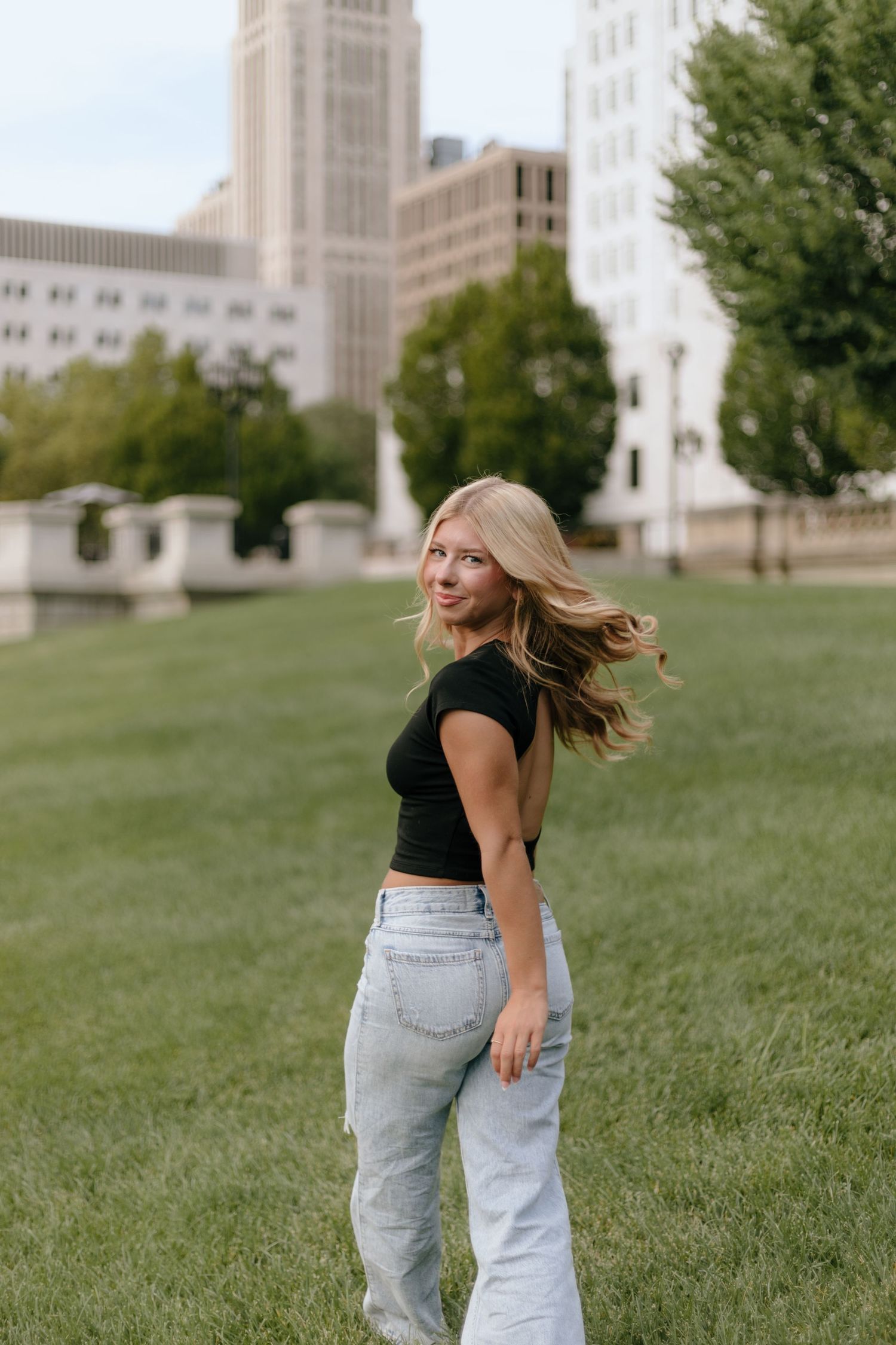 A person in a black top and light jeans dances and spins on a grassy hill with tall buildings in the background.