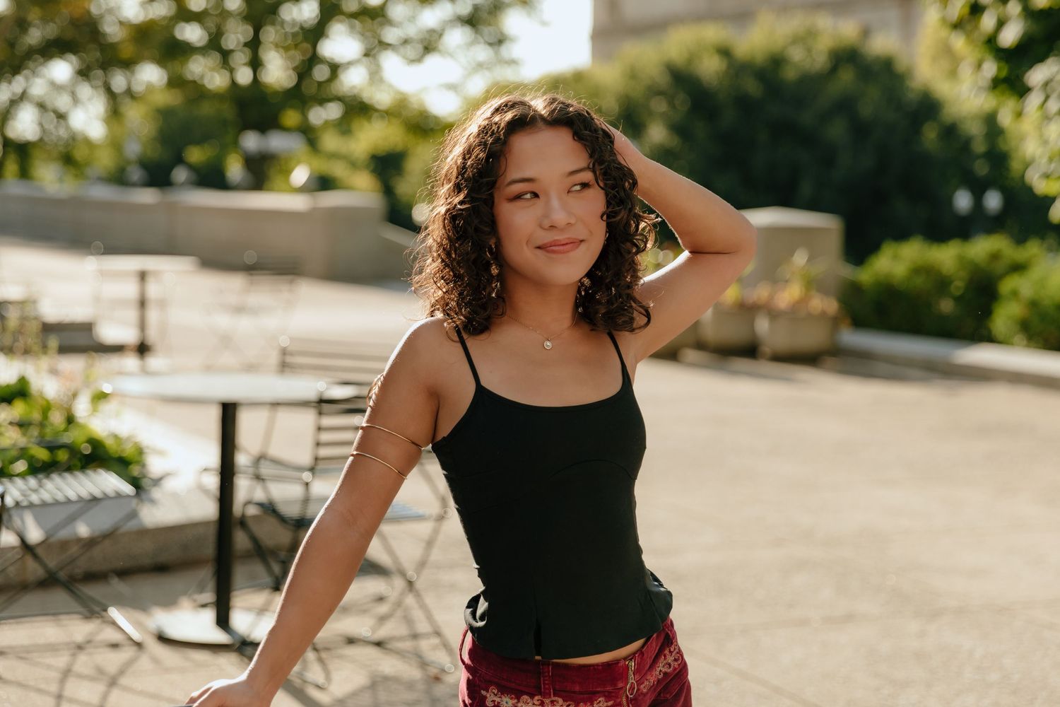 Woman in black tank top and burgundy skirt poses outdoors in sunlit urban setting.