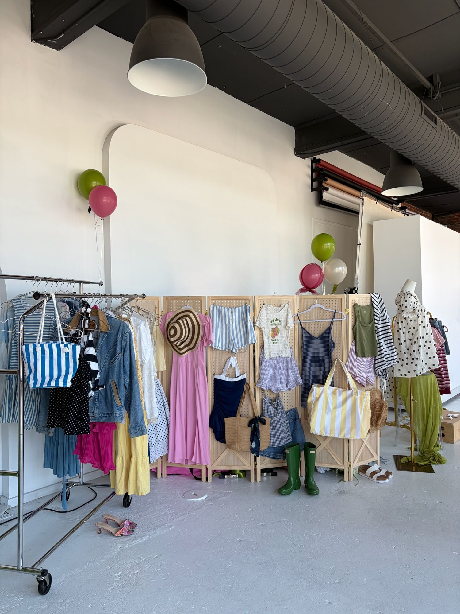 Colorful children's clothing display with balloons in a bright studio, featuring dresses, tops, and accessories on racks and wooden panels.
