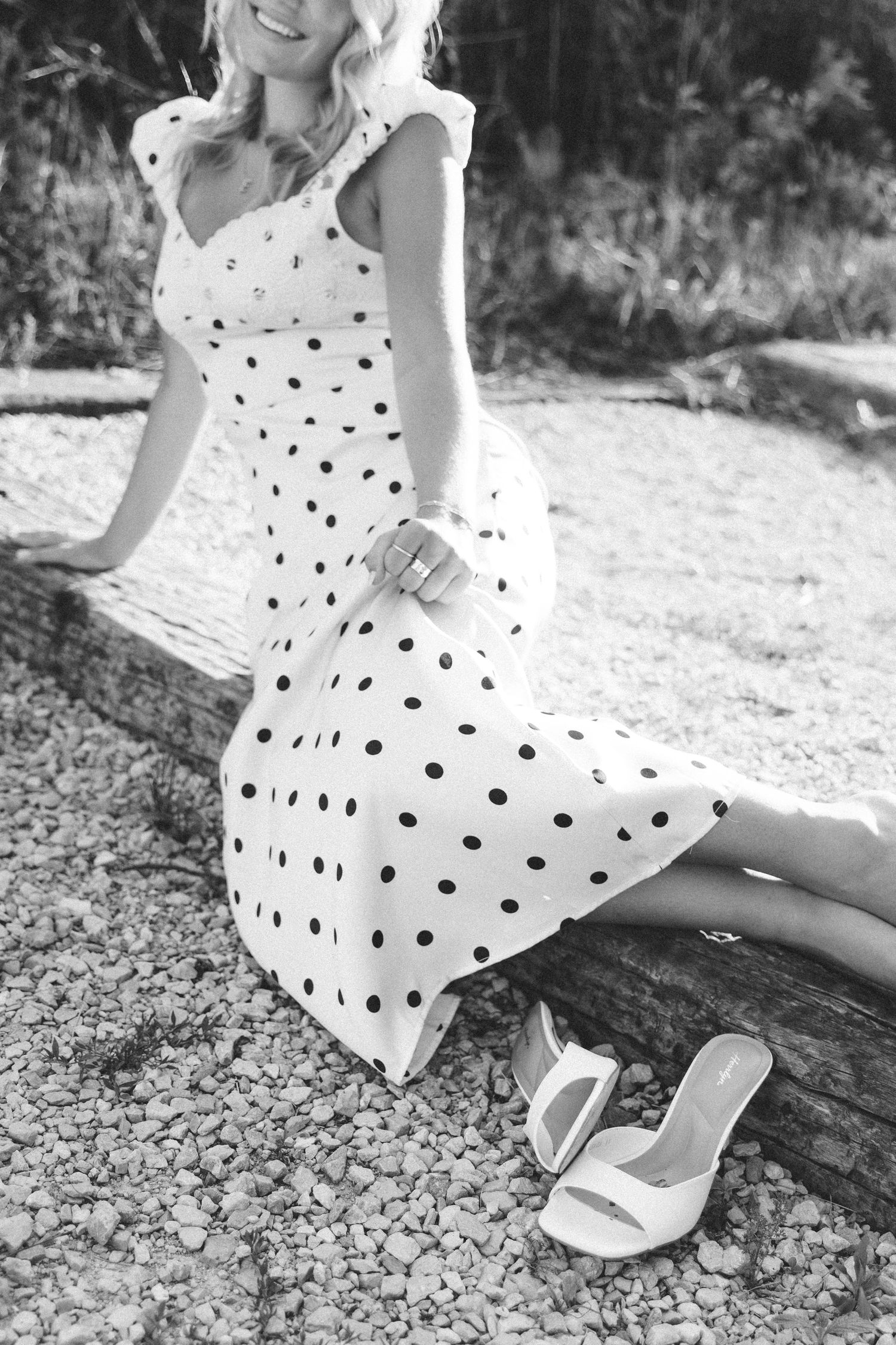 Woman in polka dot midi dress and white heels sitting outdoors on a log in a black and white photo.