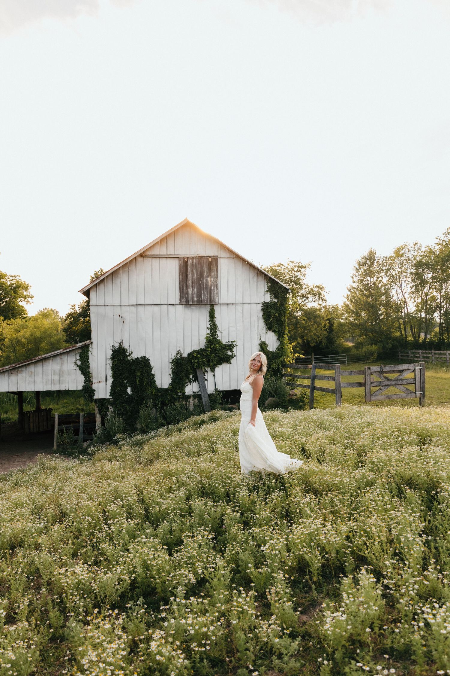 Bride in white gown standing in wildflower field before rustic white barn at golden hour.