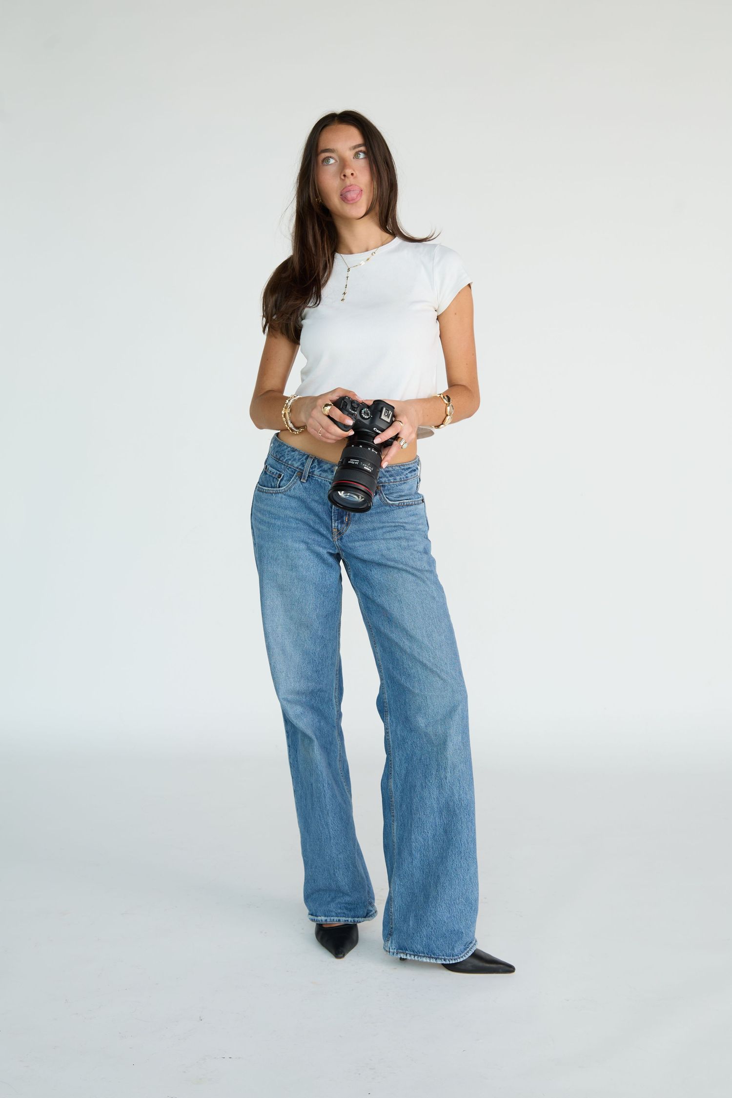 Woman in white tee and wide-leg jeans holding a DSLR camera, posing playfully in a studio setting.