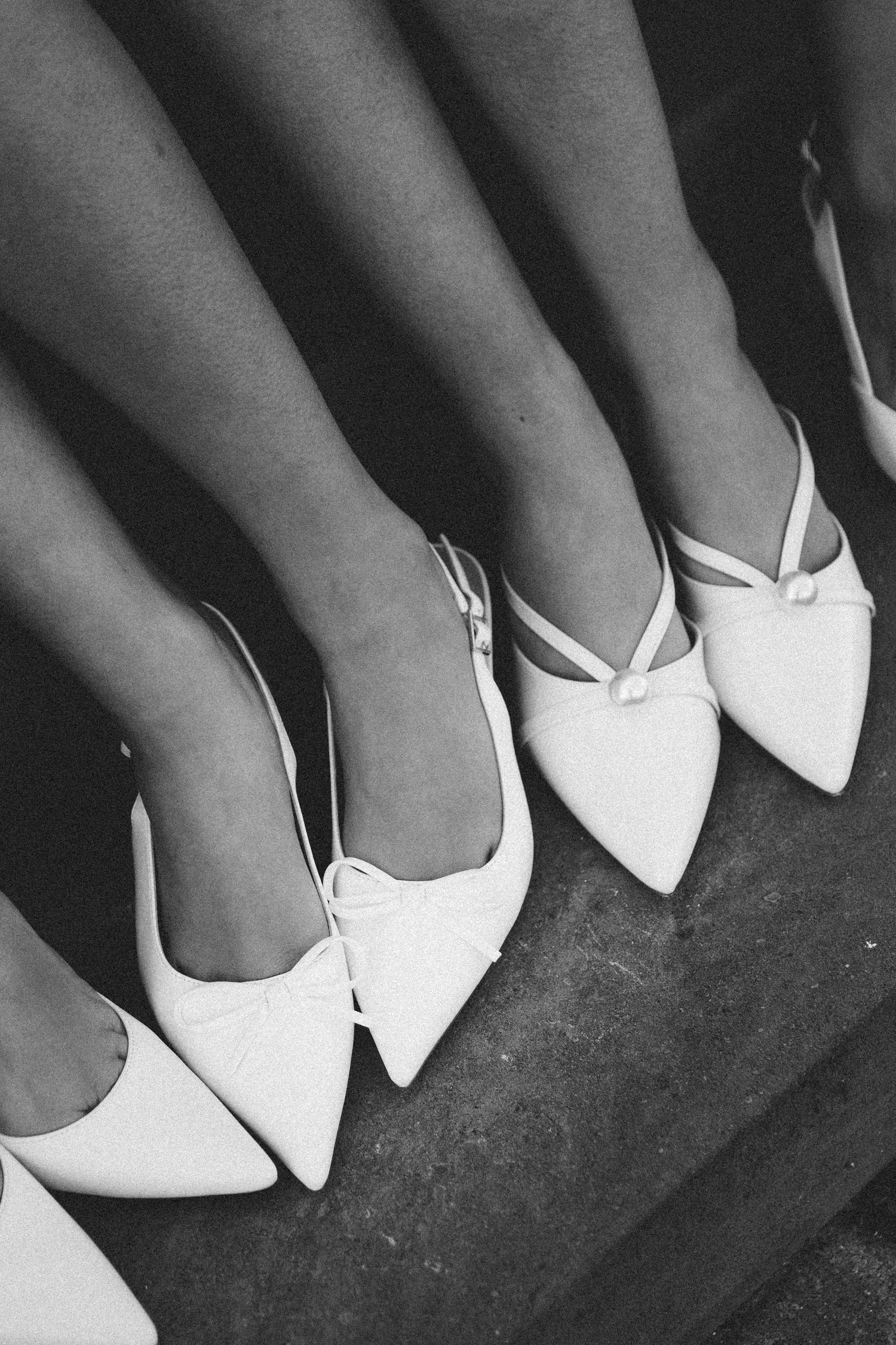 Black and white photo of multiple women wearing matching white pointed-toe slingback heels with bow detail on steps.