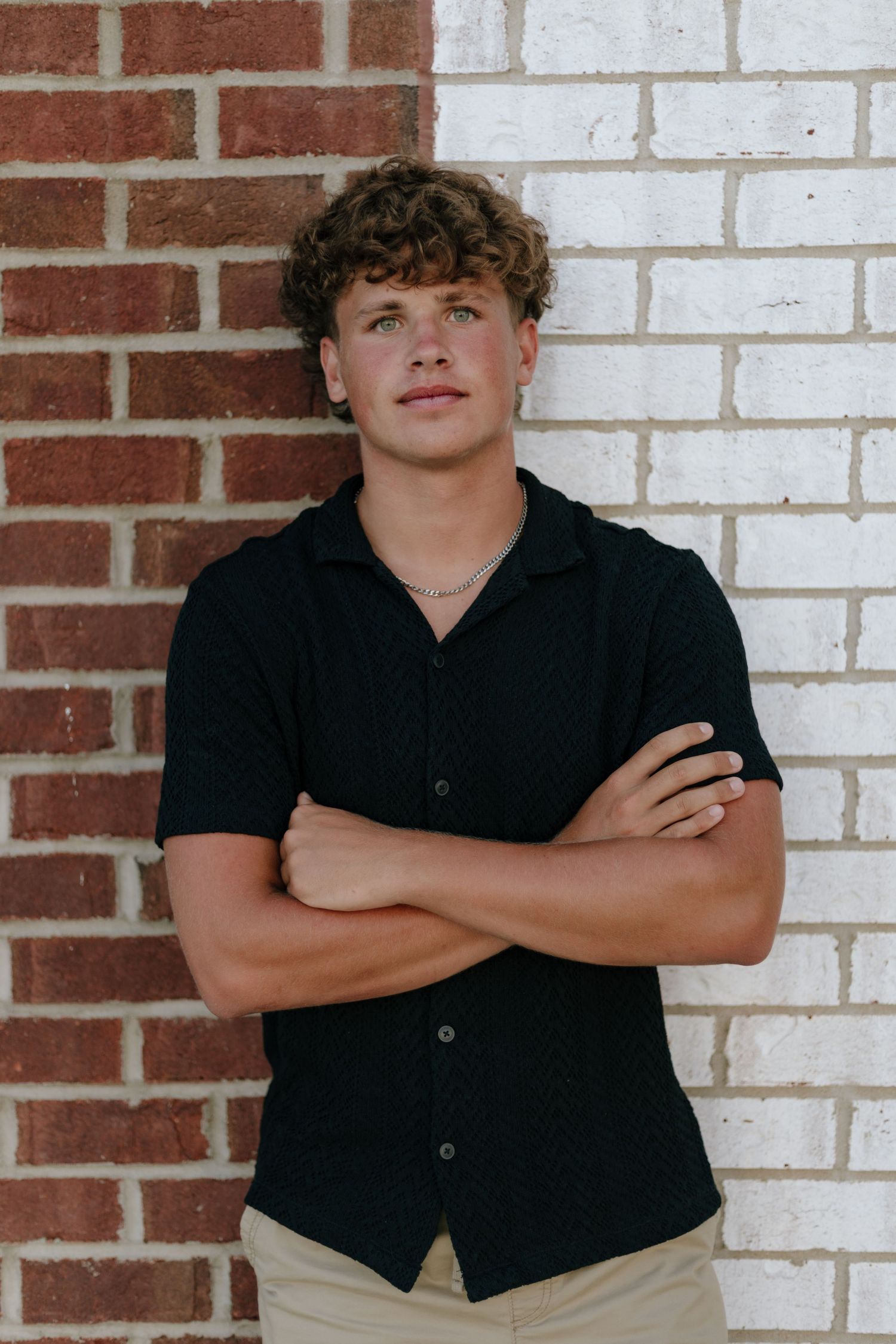 Person in black shirt poses against white brick wall with crossed arms in casual series.