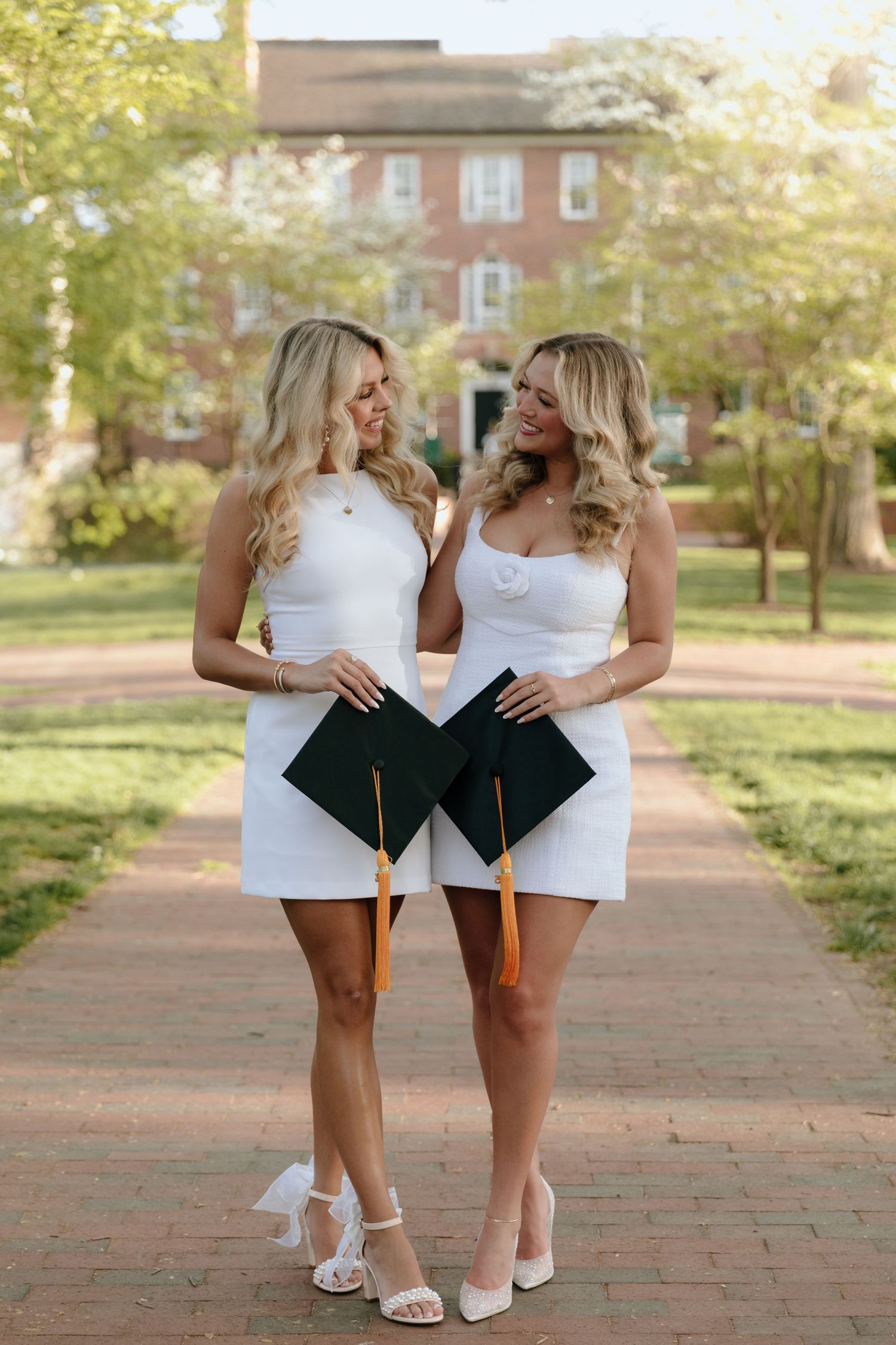 Two graduates in matching white dresses pose with their diplomas on a brick pathway with a historic building in background.