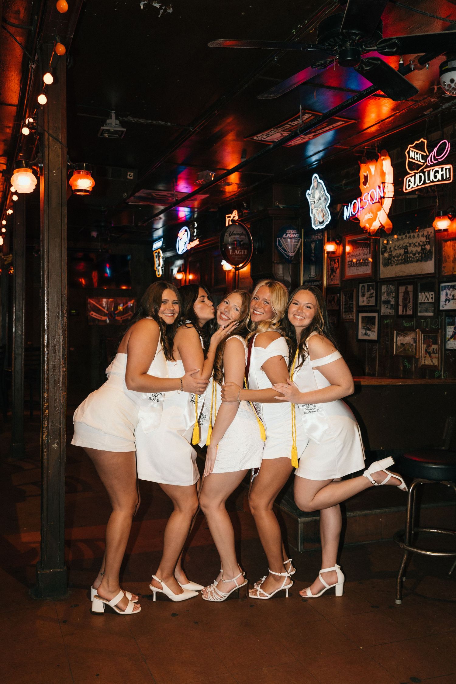 Group of women in matching white outfits posing together at a bar with neon signs.