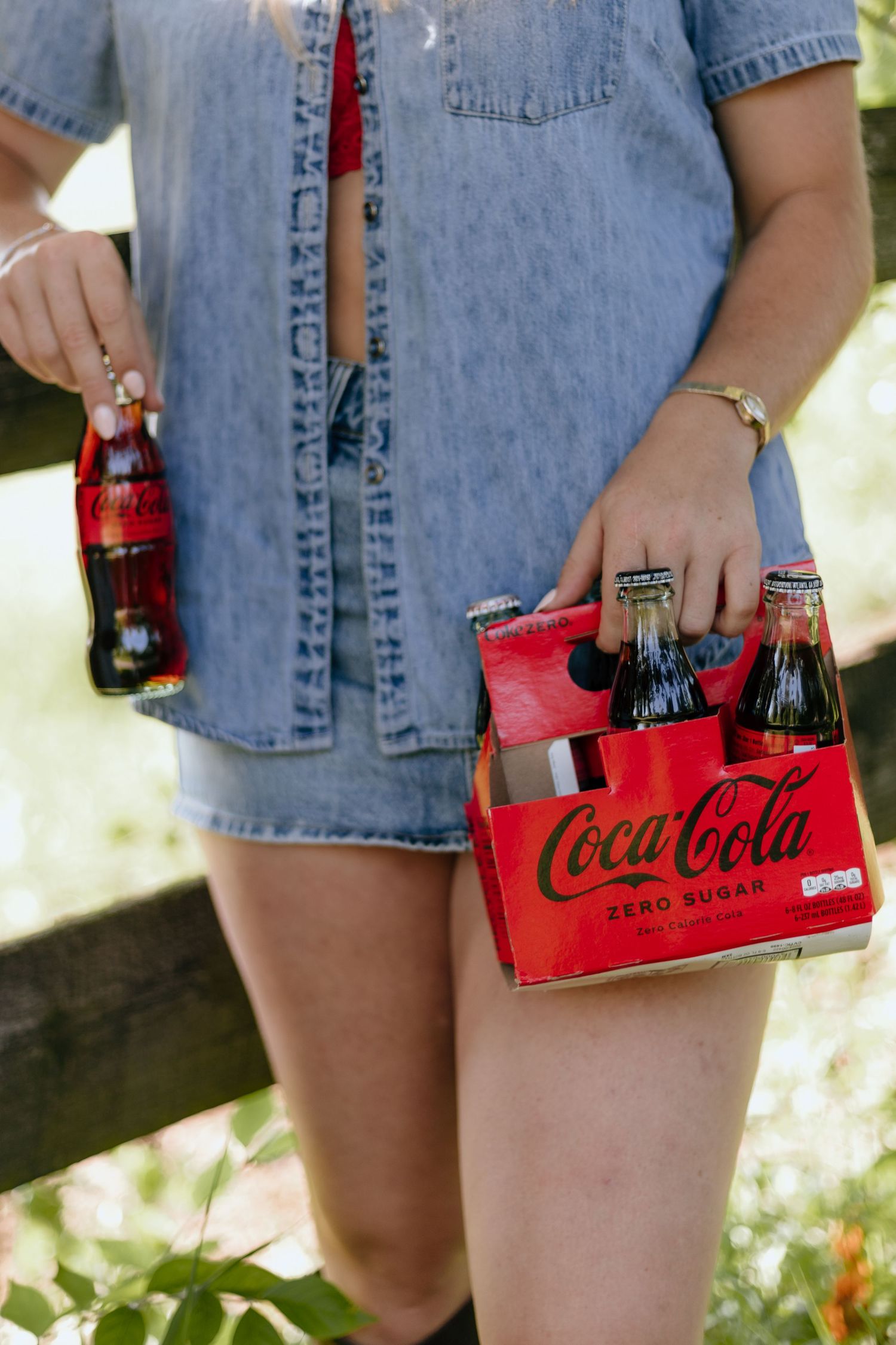 Person in denim shirt holding classic red Coca-Cola bottles and carrier on a sunny day outdoors.