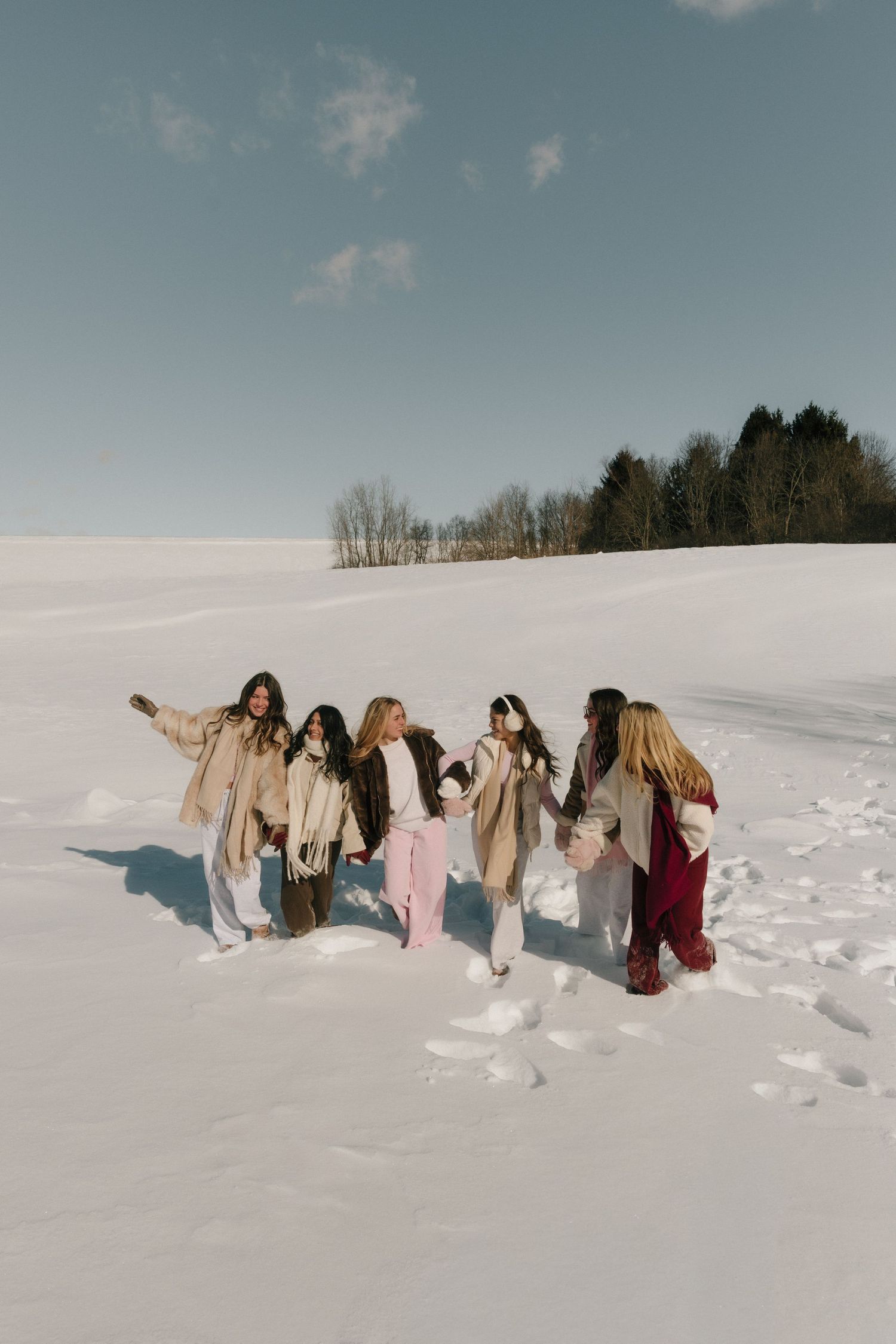 Group of young women walking together in a snowy winter landscape on a sunny day, wearing cozy neutral-toned coats.