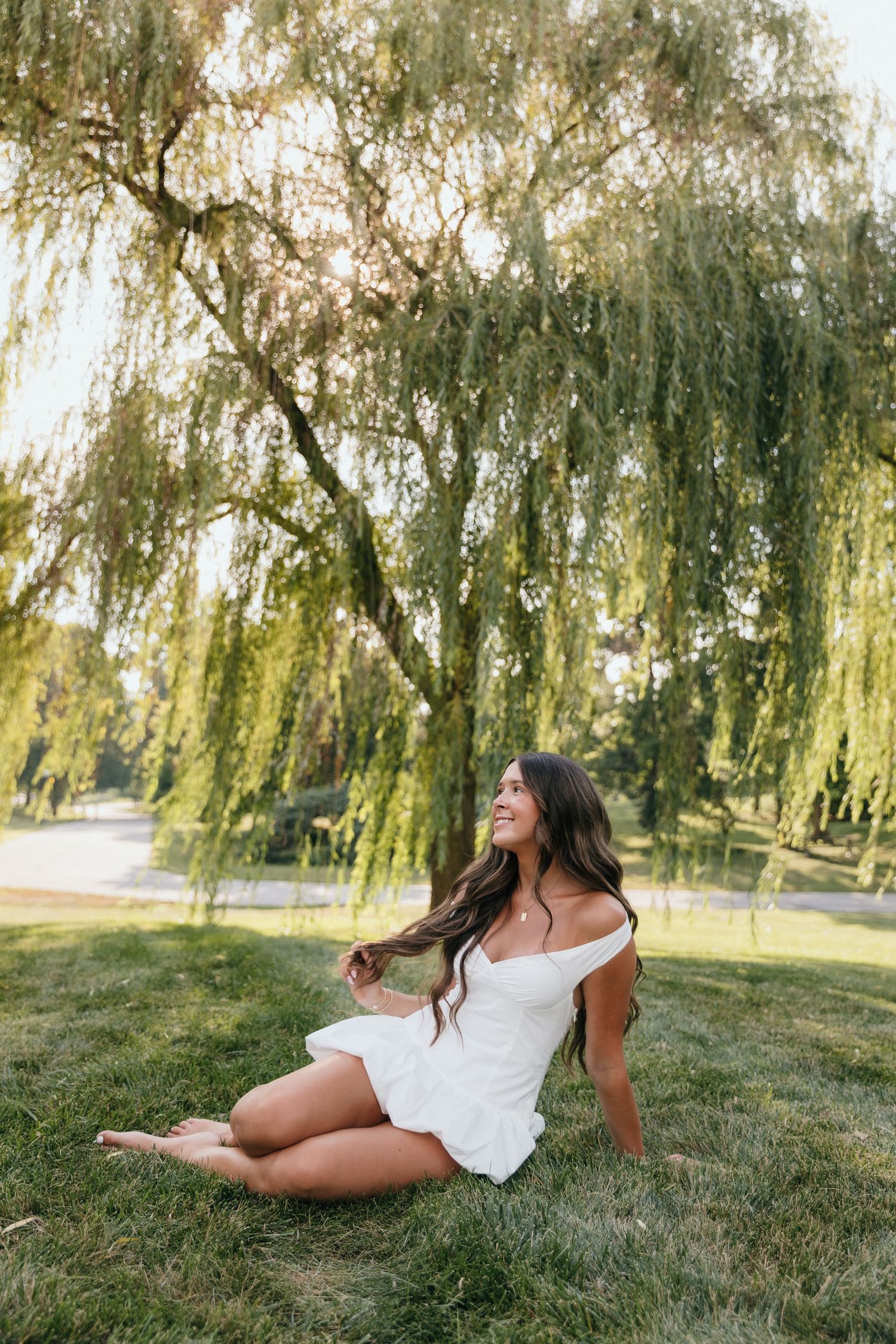 Young woman in white dress sitting on grass under a weeping willow tree, smiling in golden sunlight.