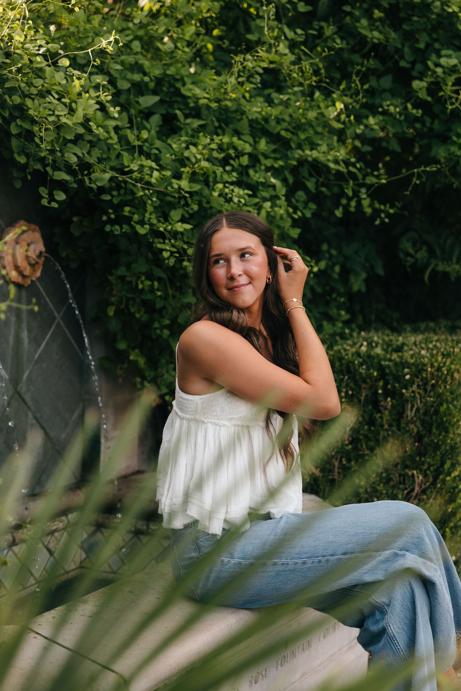 Young woman in white top and jeans sitting by a garden fountain surrounded by lush green foliage.