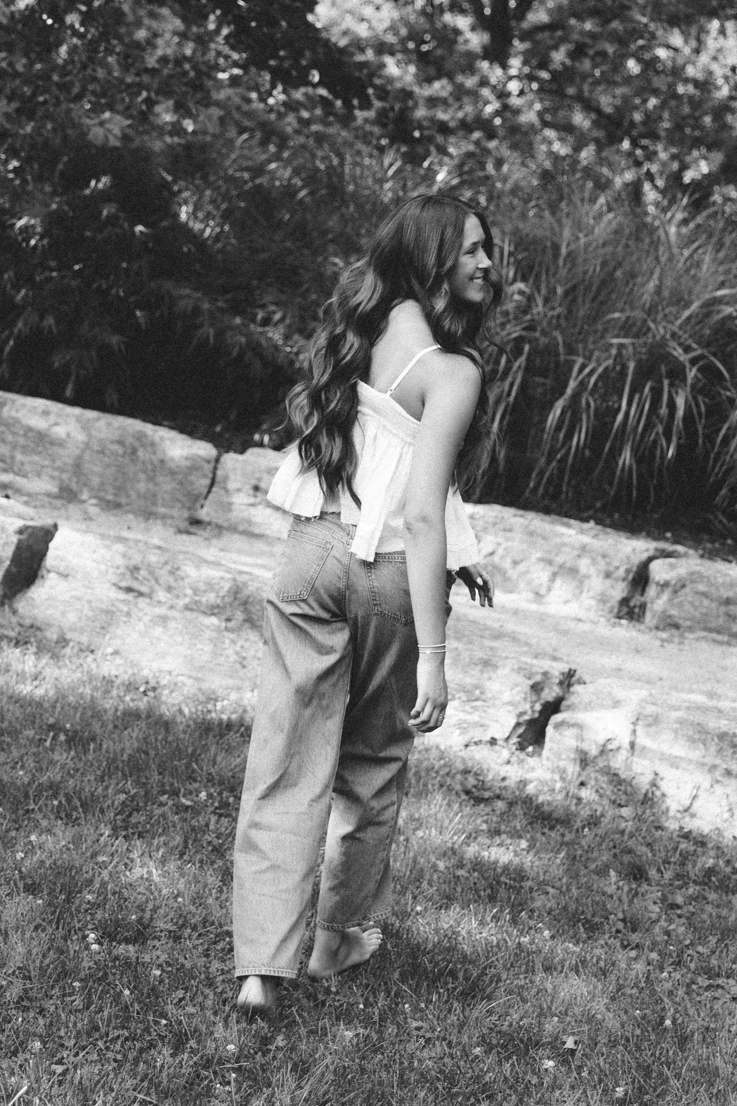 Black and white photo of a young woman with long wavy hair wearing wide-leg pants and a white top in a park.