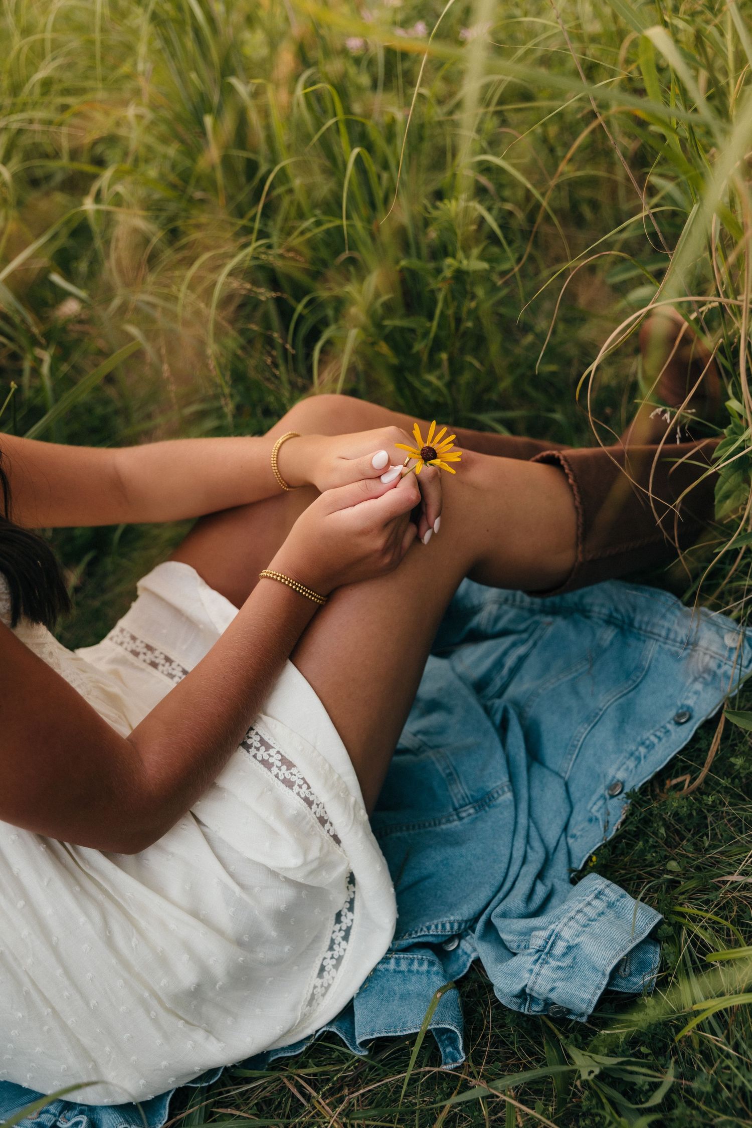 Woman sitting in a grassy field holding a small yellow wildflower, wearing a white top and denim skirt.
