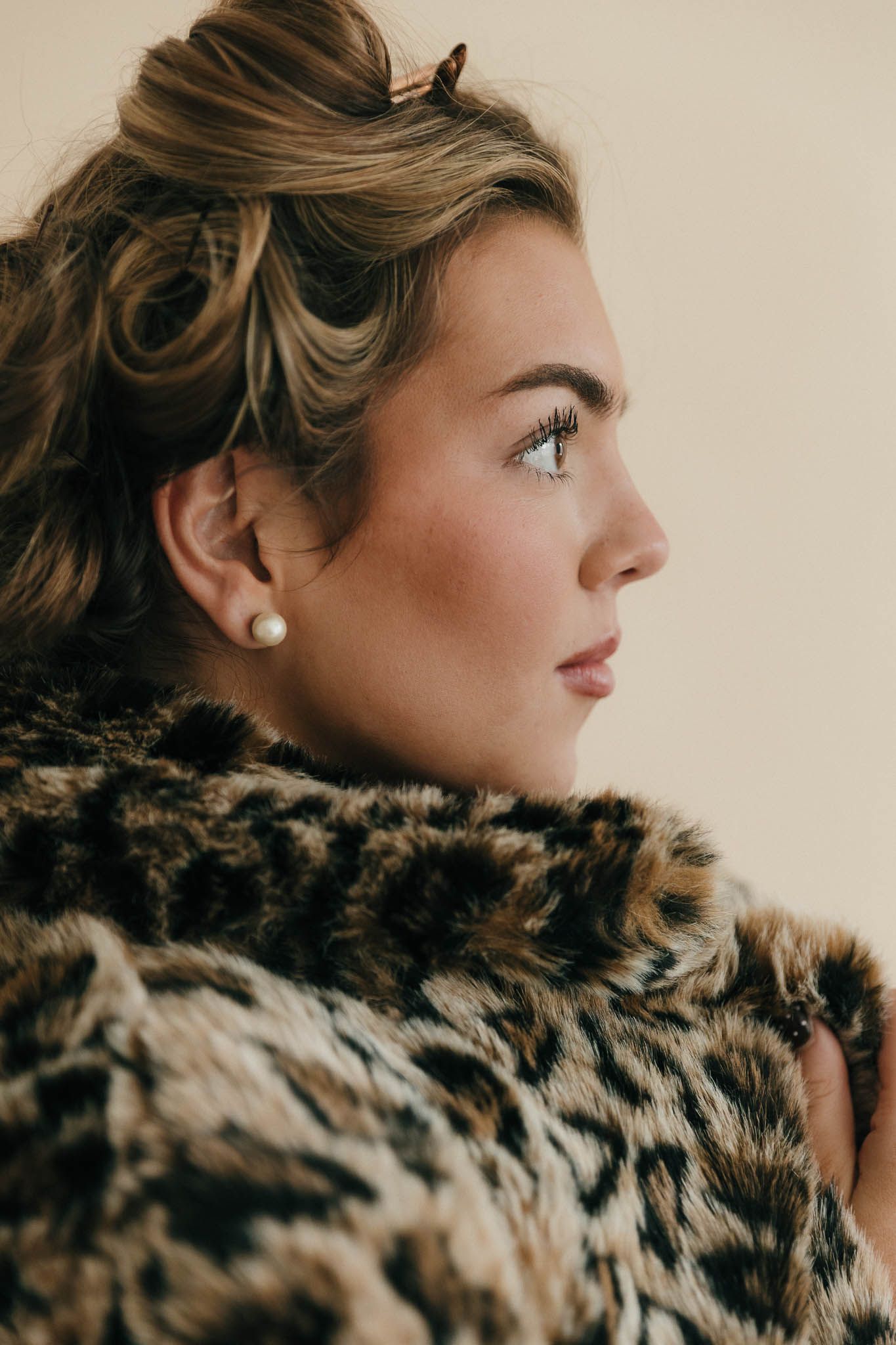 Side profile portrait against beige background wearing fur collar coat and pearl earring in warm tones.