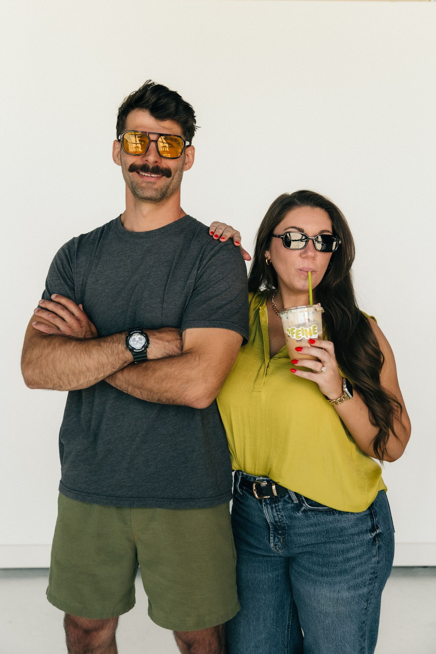 A man and woman wearing novelty sunglasses pose together, she sips a drink while he stands with arms crossed.