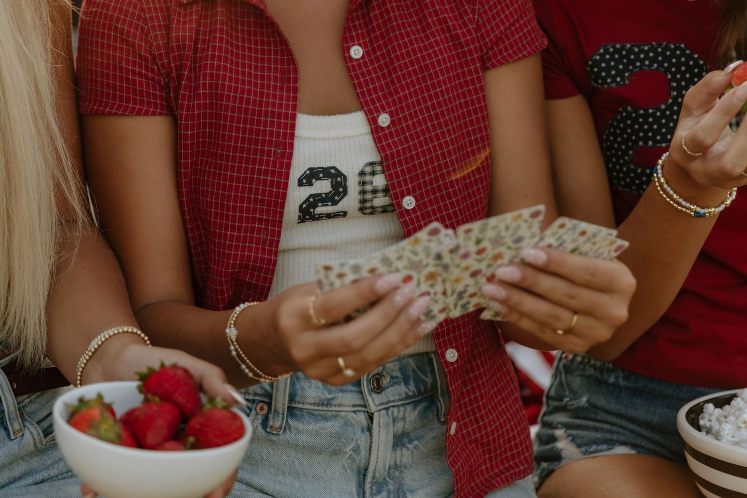 Two women sitting together holding playing cards with bowls of fresh strawberries and snacks nearby.
