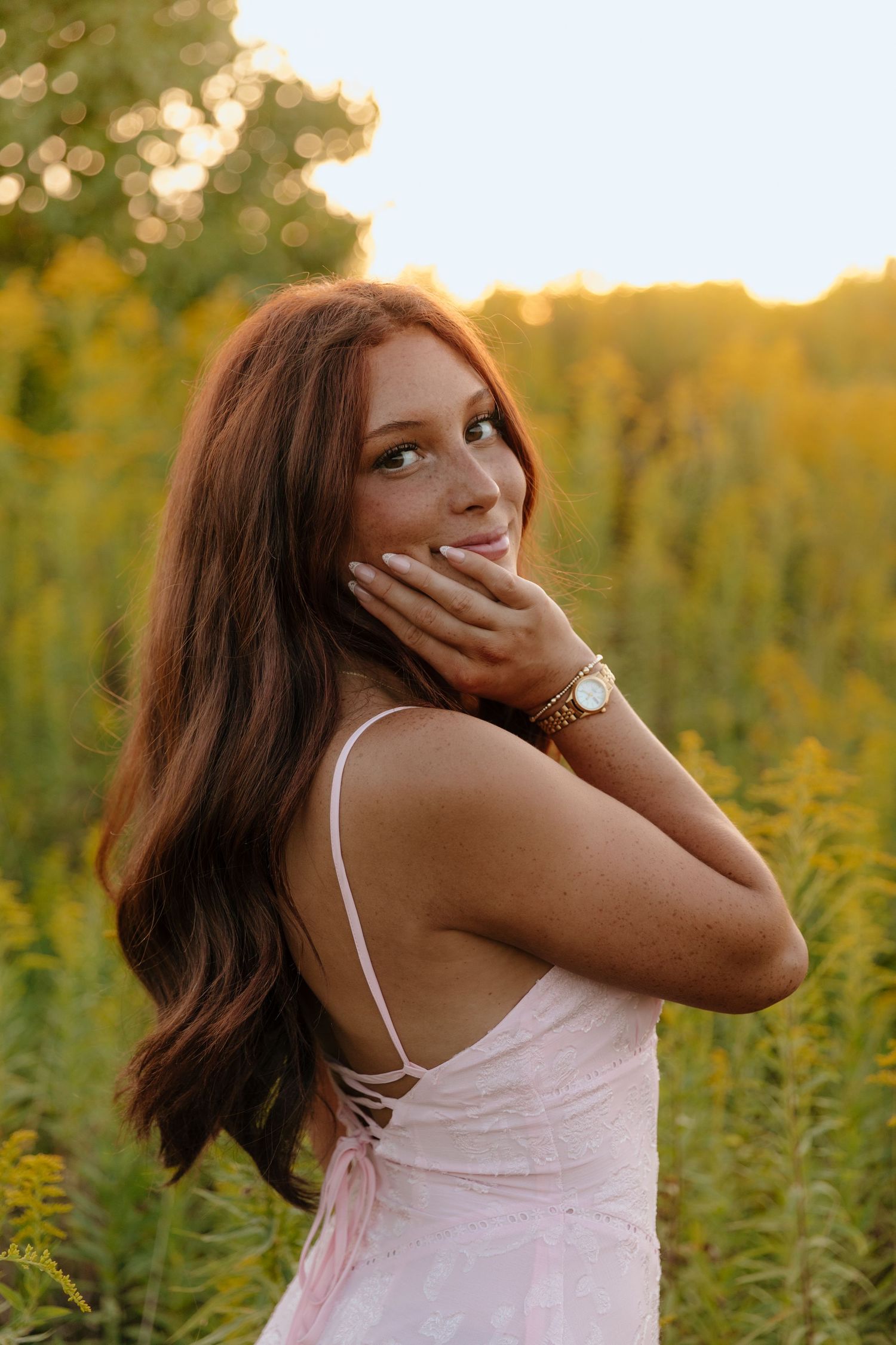 A person in a light pink camisole poses playfully in a field of wildflowers during golden hour sunset.