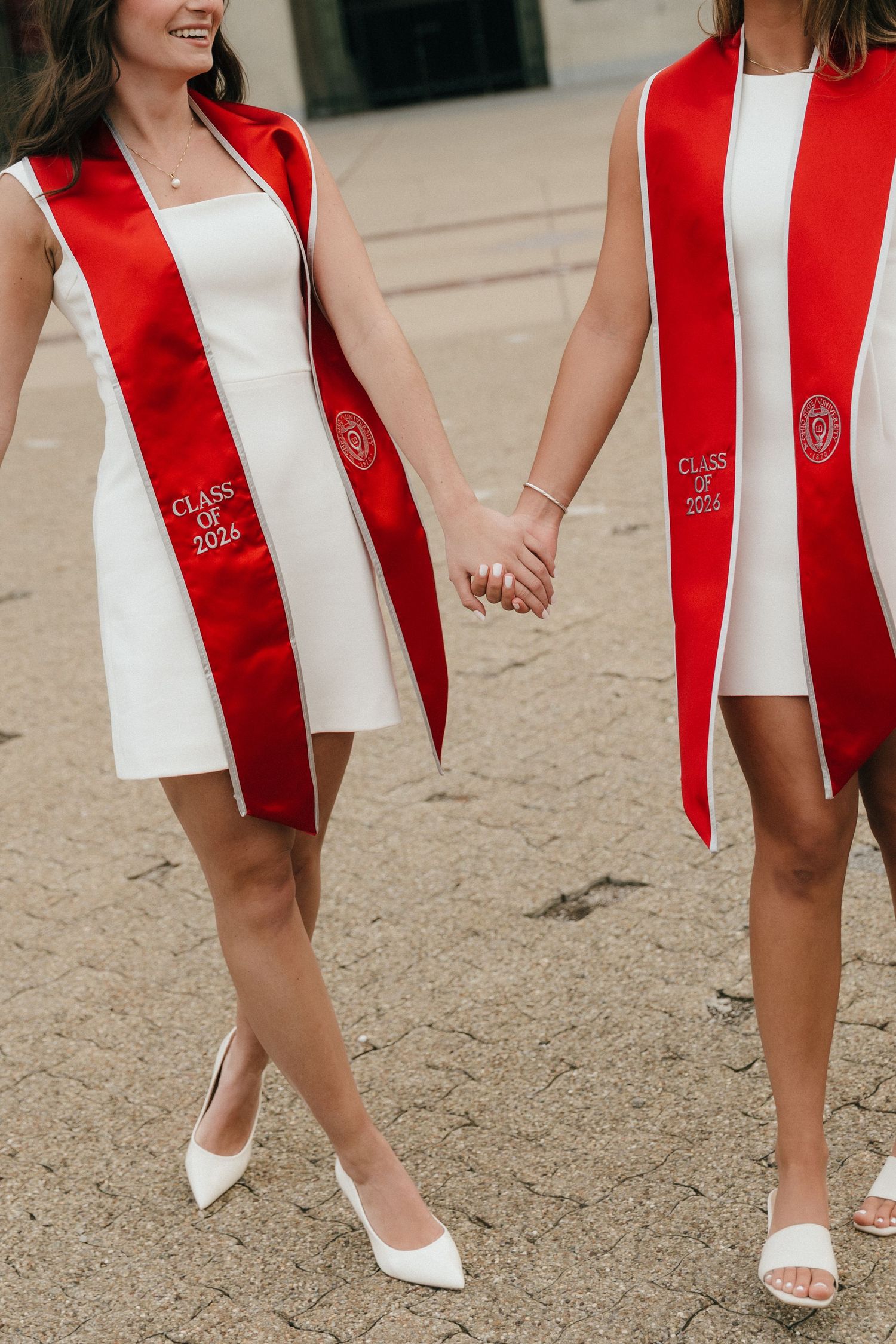 Two graduates in white dresses and red Class of 2025 sashes hold hands outdoors.