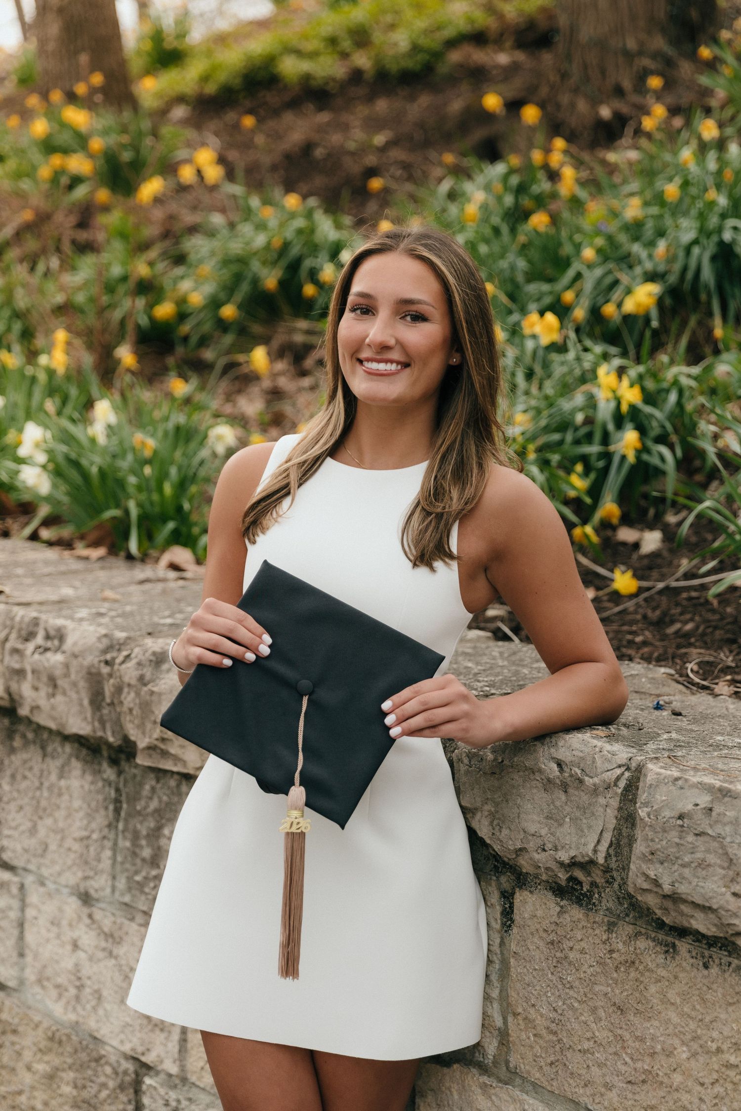 Smiling graduate in white dress holds black cap near spring daffodils on stone wall.