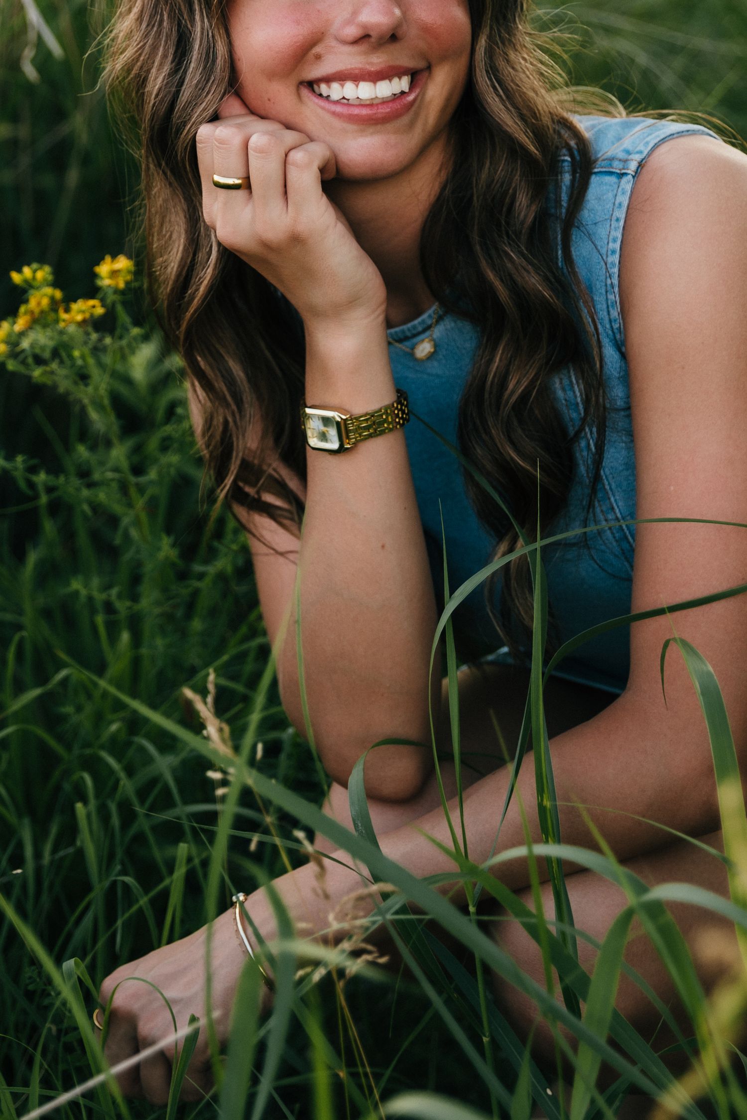 Smiling young woman sitting in green grass field wearing denim vest, gold watch, and ring, with yellow wildflowers.