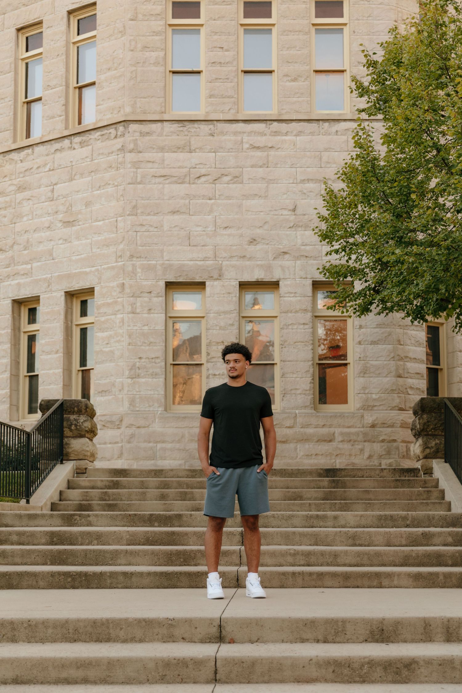Young man in black t-shirt and gray shorts standing on stone steps outside a large limestone building.