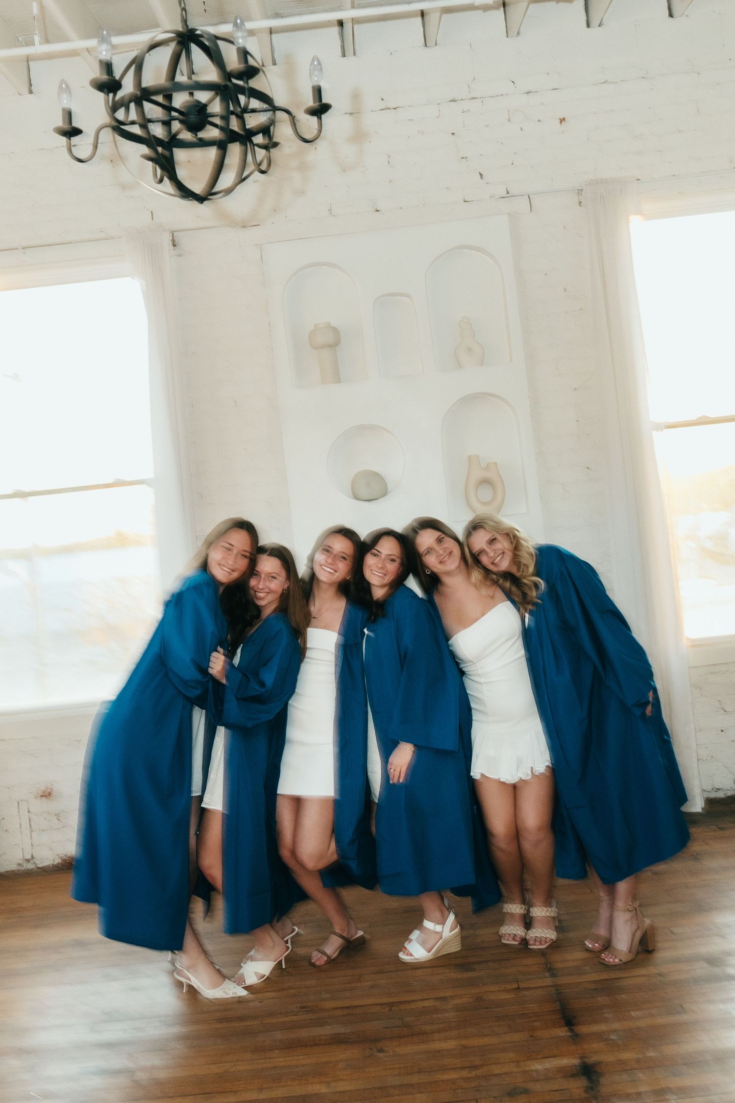 Six women posing together in blue robes and white dresses in a bright white room with hardwood floors.