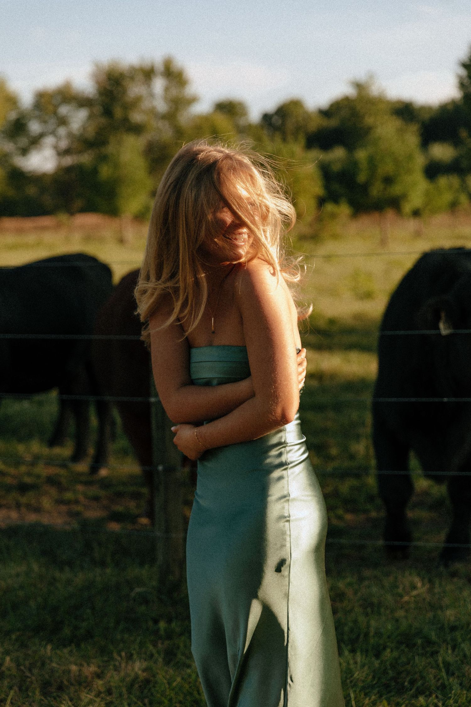 A person in a turquoise dress stands in a sunlit field at golden hour with horses grazing in the background.