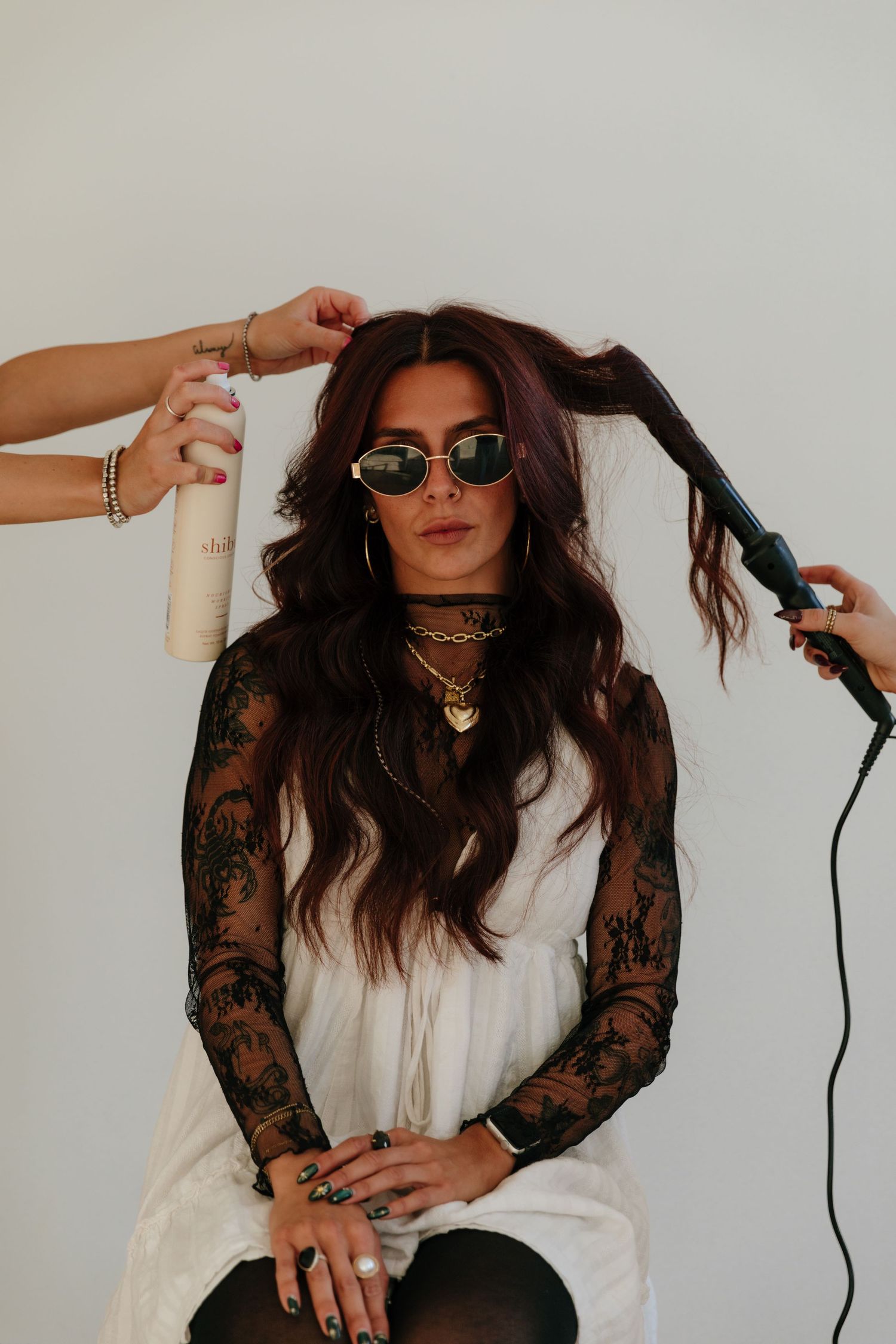 Person in black lace and white top styling hair with curling iron while wearing sunglasses in a photo series.