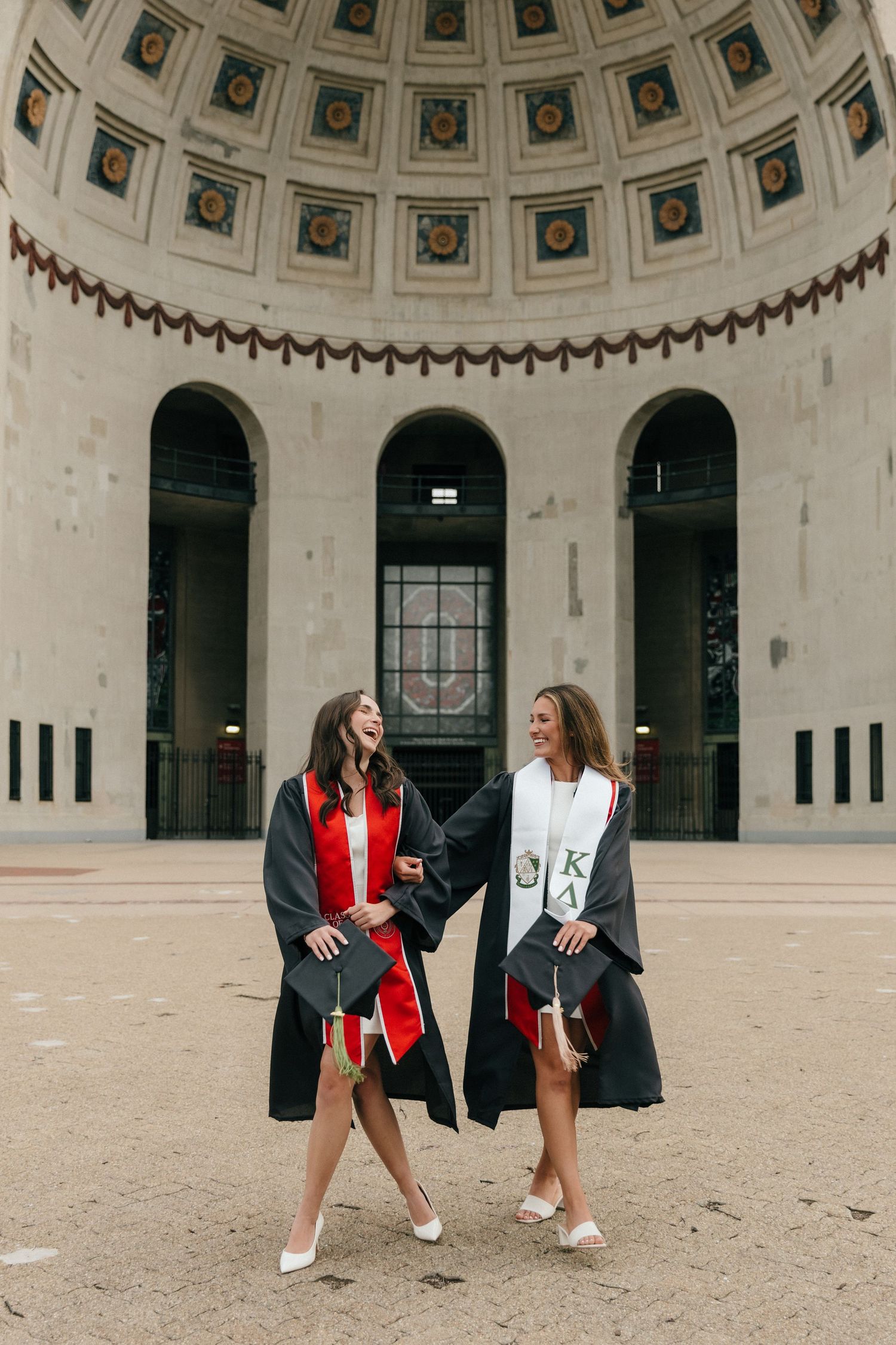 Two female graduates in black gowns with stoles laugh together outside a domed university building.