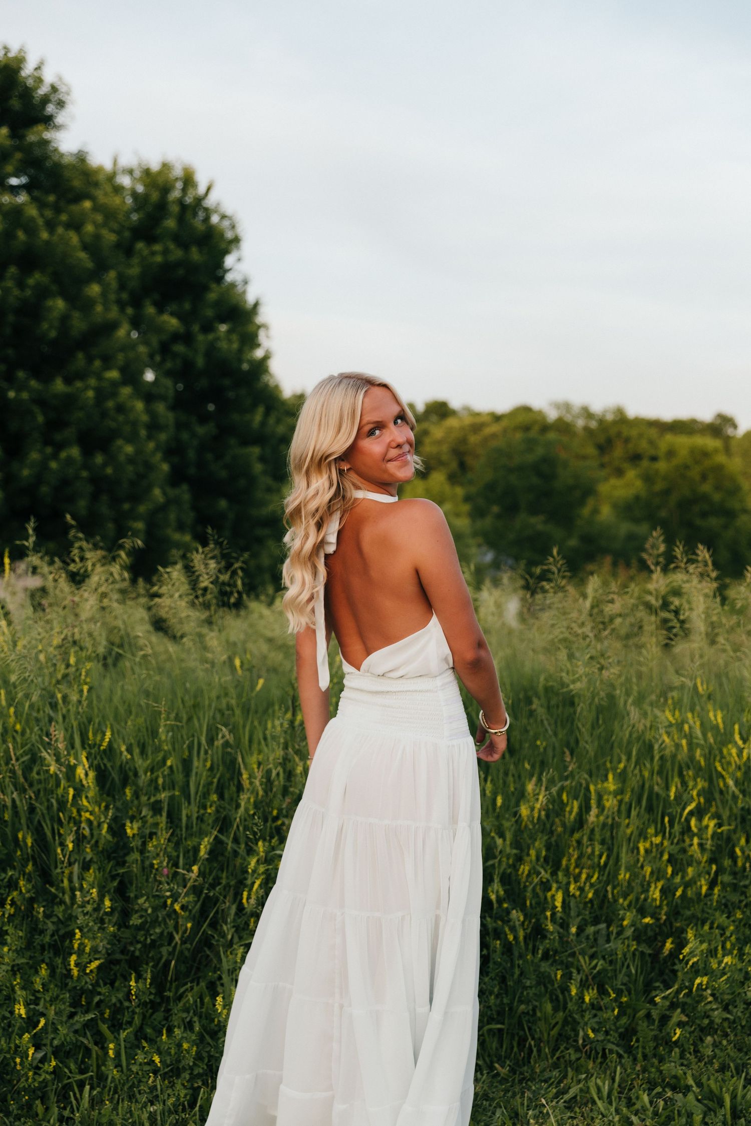 Blonde woman in white backless halter maxi dress standing in green field, smiling over shoulder at golden hour.