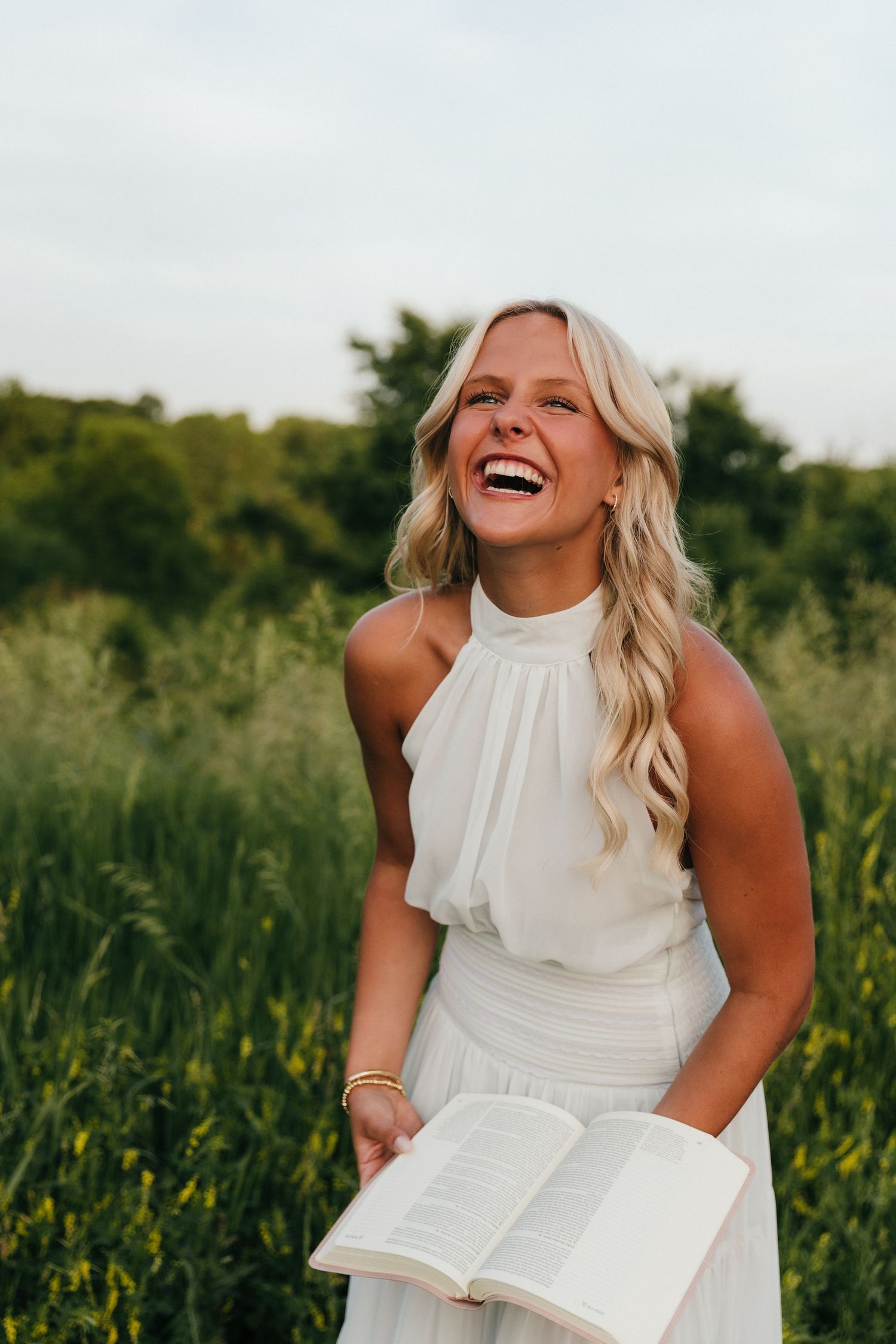 Laughing blonde woman in white dress holding open book outdoors in green field.